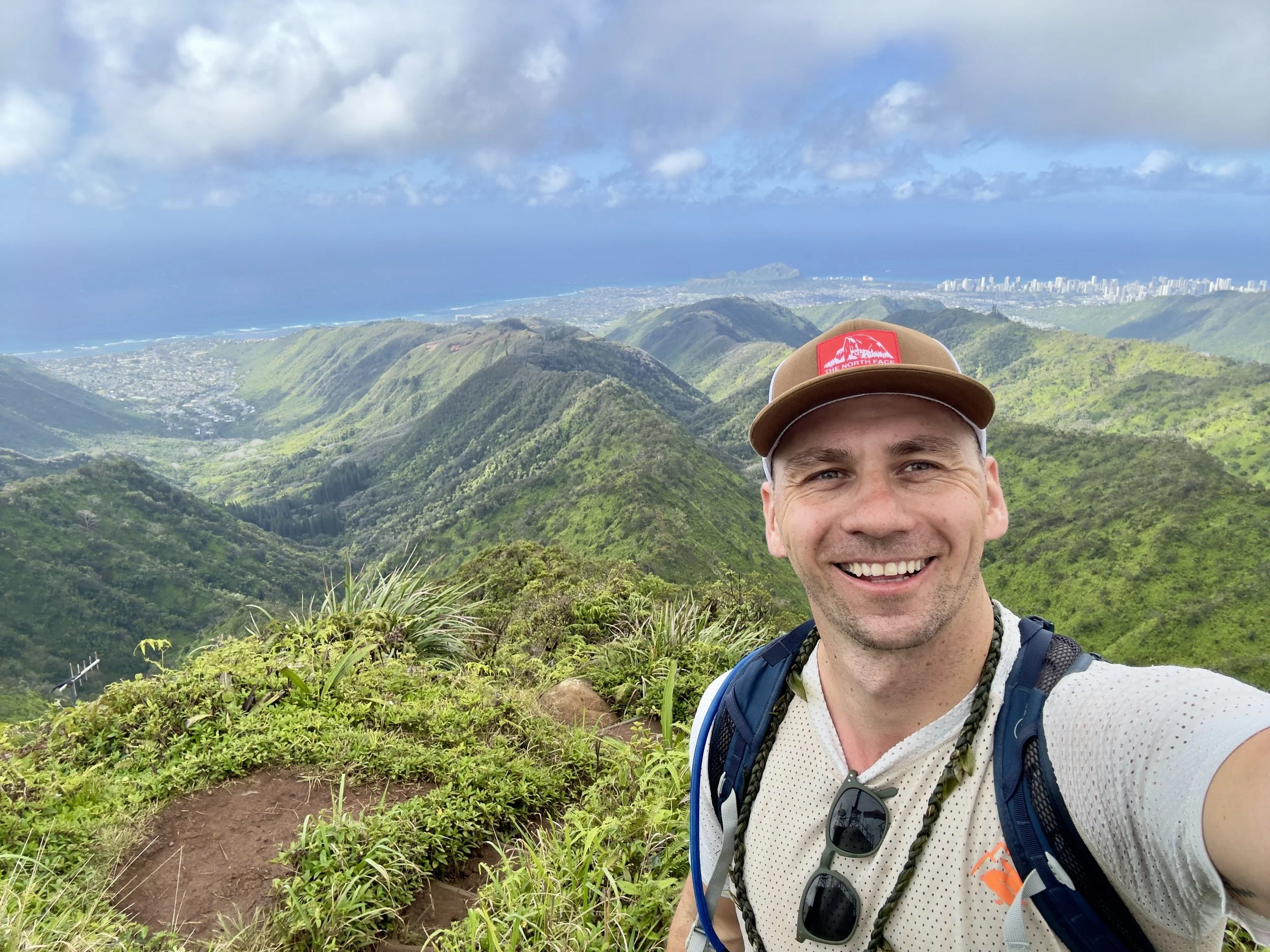Smiling man taking a selfie on a mountain trail with lush green mountains, a city year, and an ocean in the background.