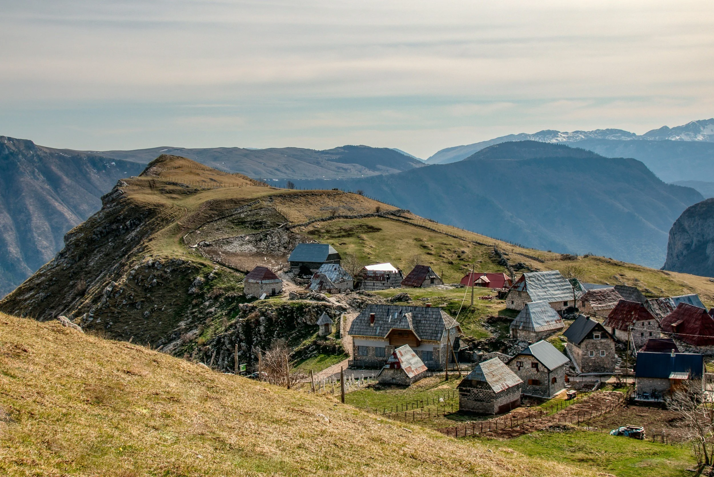 A small rural village with houses and barns on a hillside, surrounded by mountains and rolling terrain.
