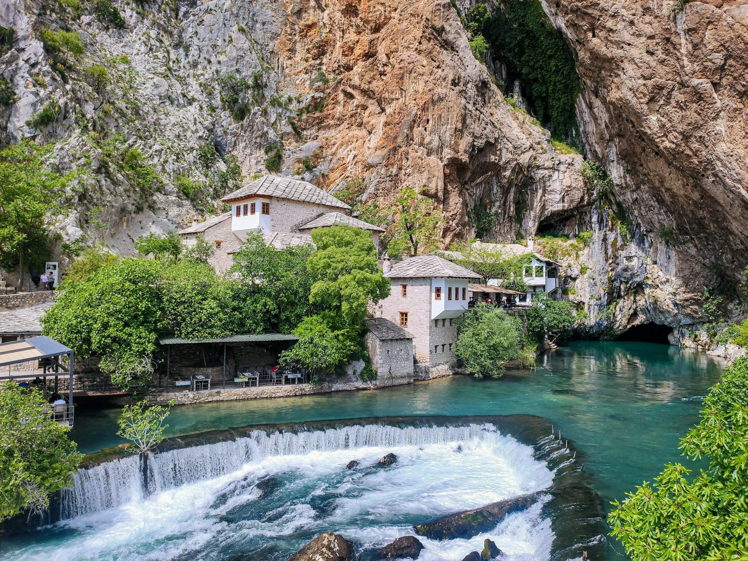 A scenic view of a small village with white buildings built into a rocky cliffside beside a flowing river, with lush green trees and a rock cave visible in the background.