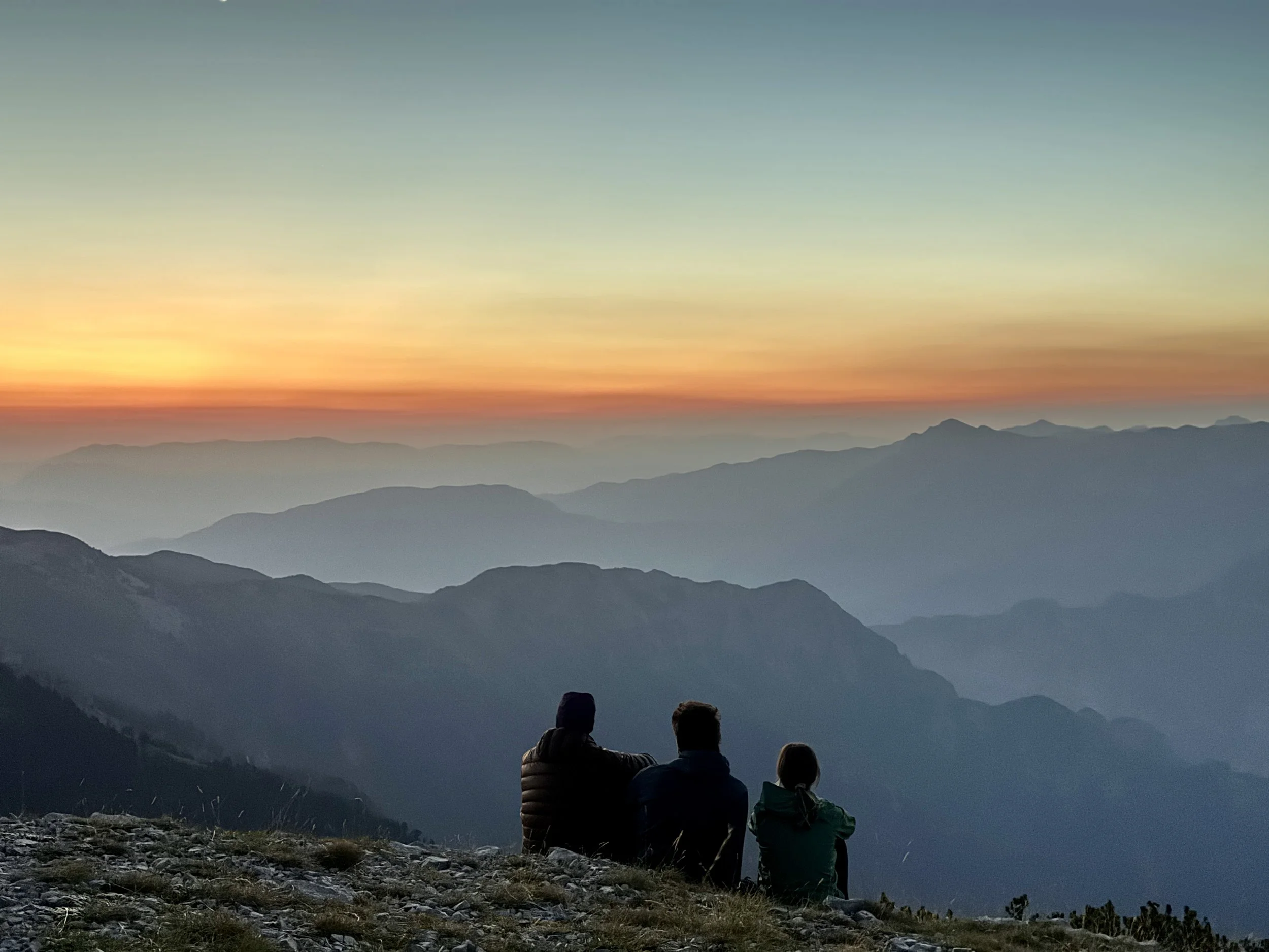 Three people sitting on a rocky hillside watching a sunset over layered blue mountains.