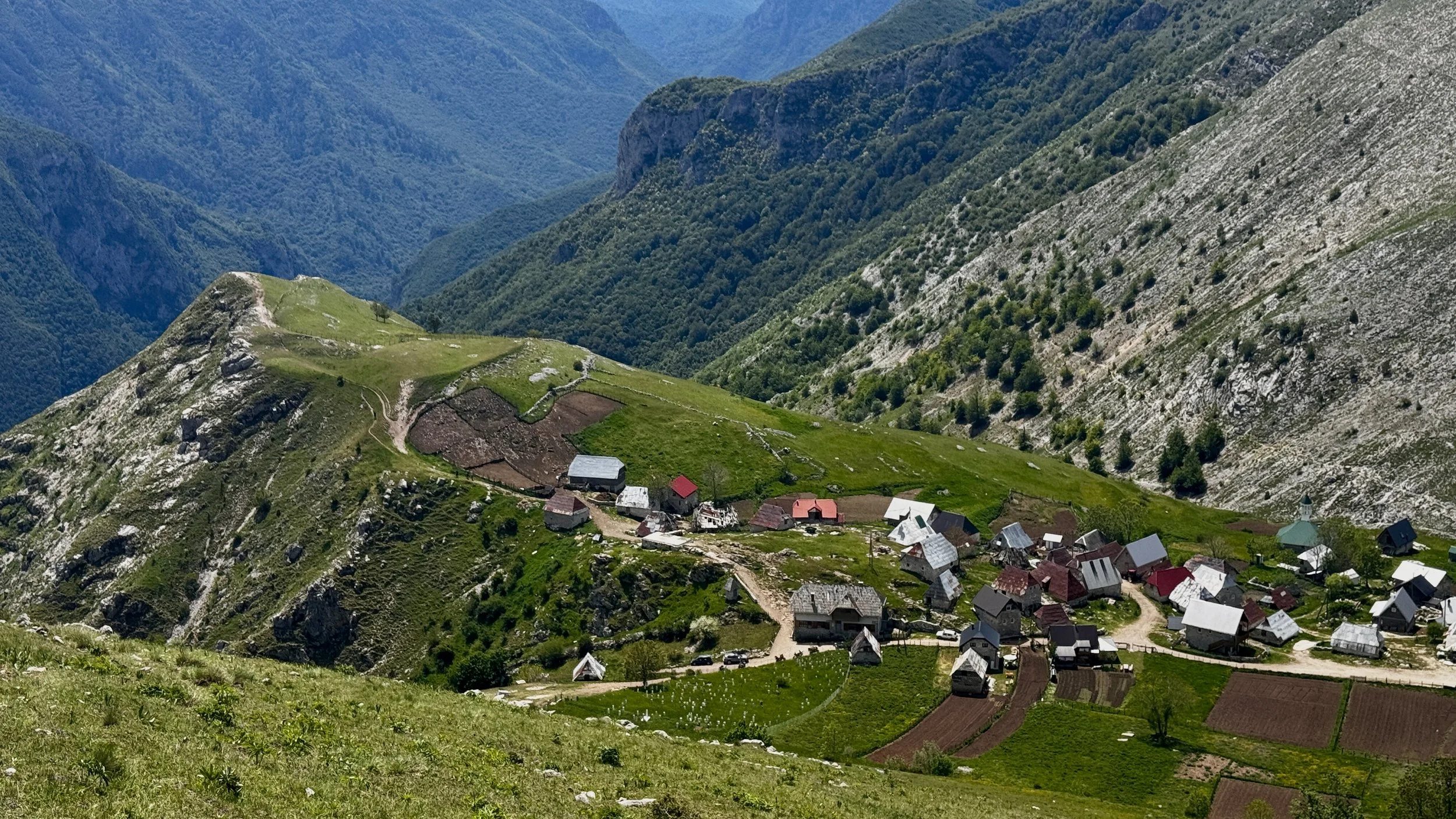 A small mountain village with houses and farmland surrounded by green hills and steep rocky mountains in the background.