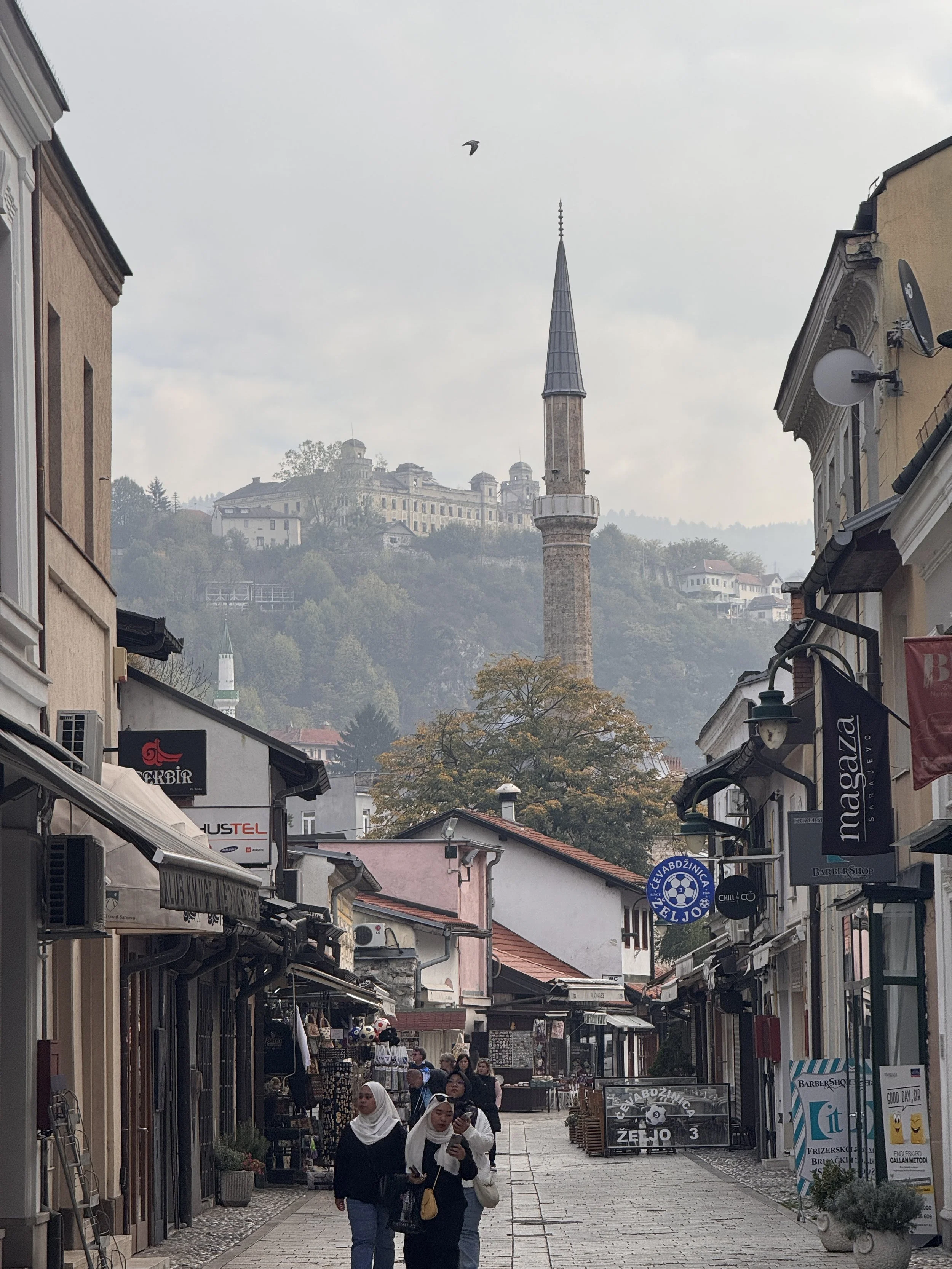 A narrow street in Sarajevo with shops and pedestrians, with a mosque minaret and a historic building on a hill in the background.