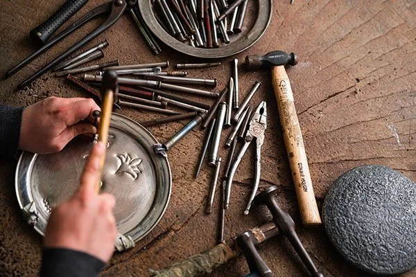 A metal craftsman's workspace with various tools, including hammers, pliers, and chisels, and a person holding a small metal object under a magnifying glass.