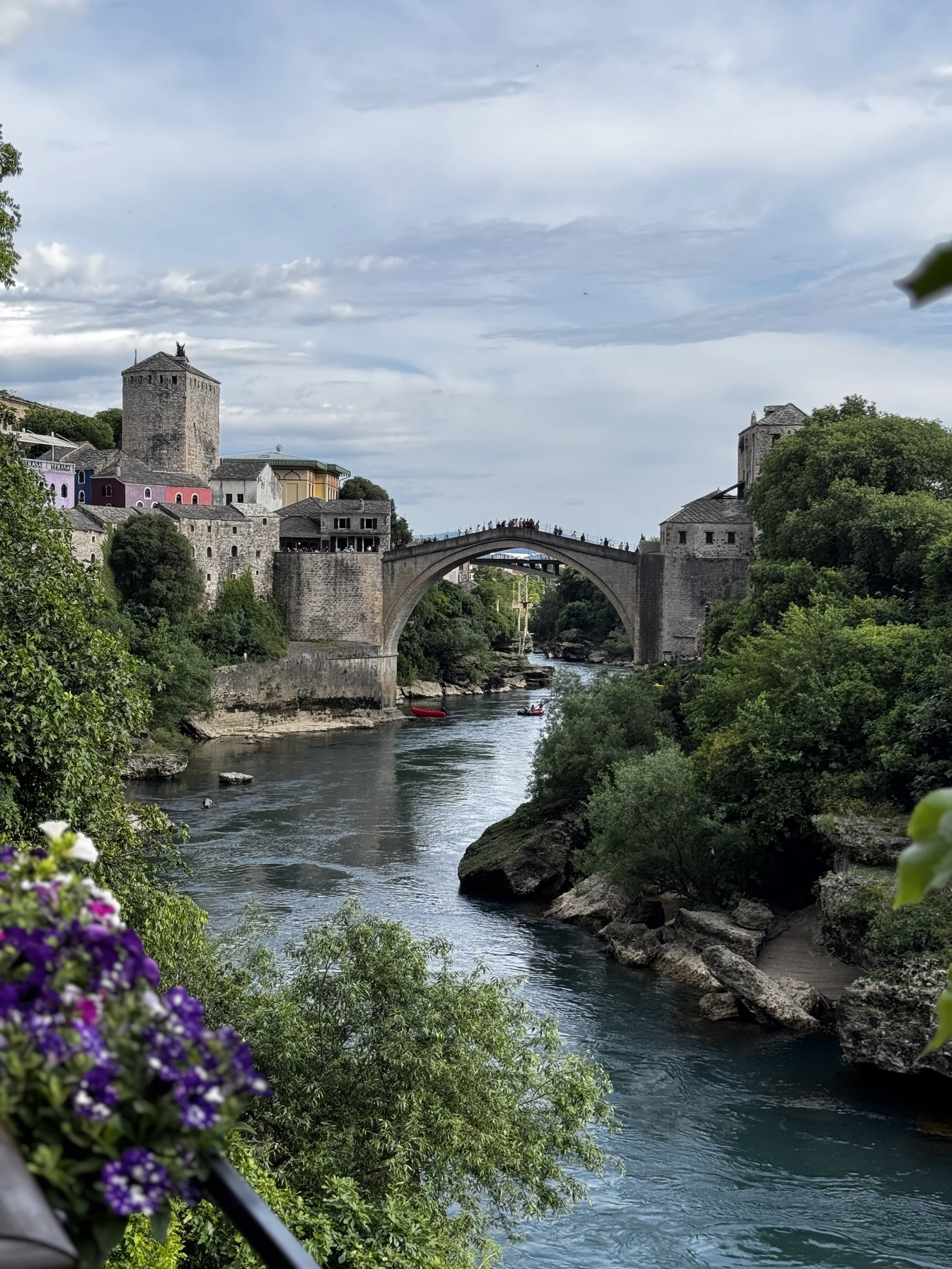 A scenic view of the old town of Mostar with historic stone buildings and a famous arched bridge over the Neretva river, surrounded by lush green trees and boats on the water.