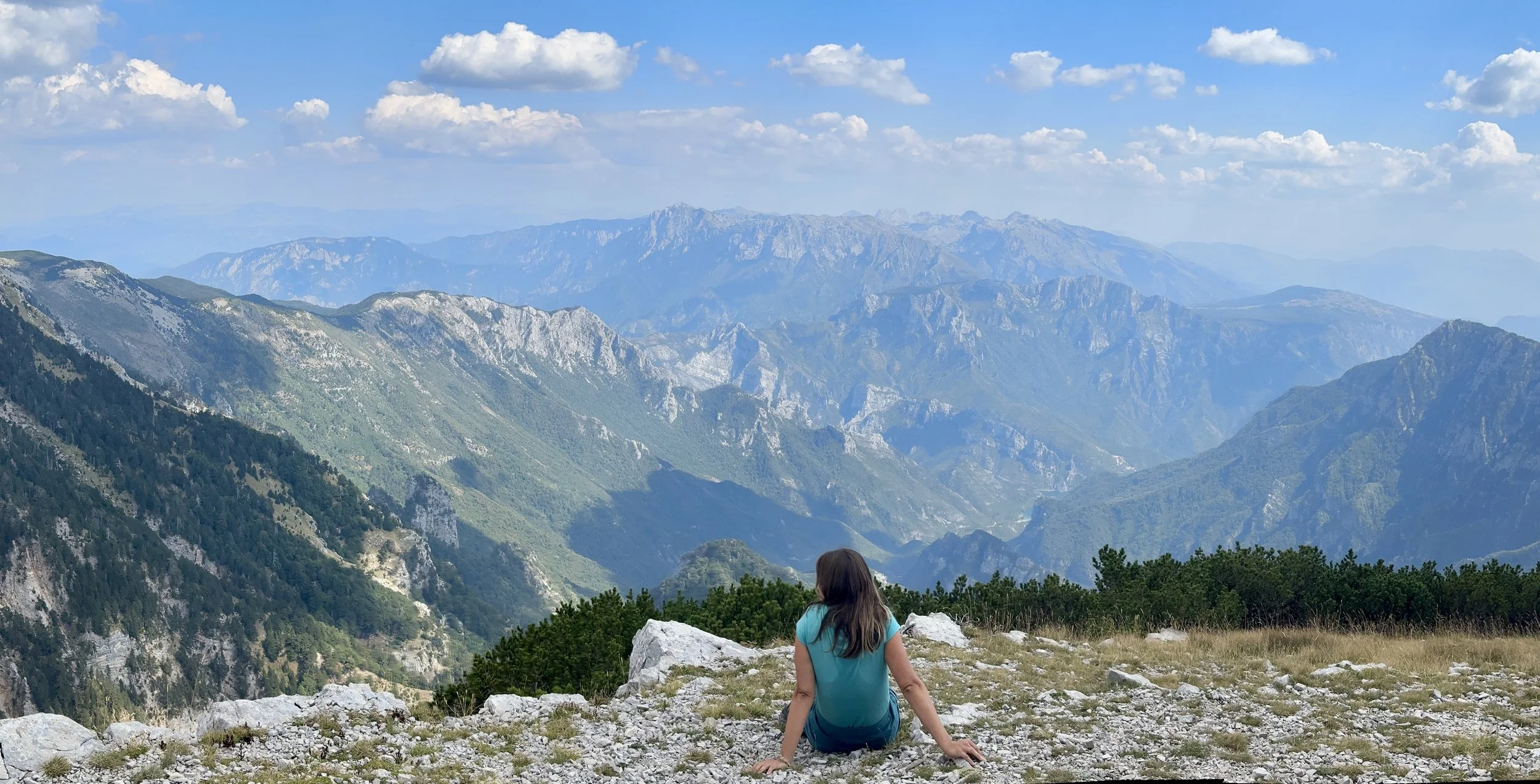 A woman sitting on a rocky mountain ledge overlooking a deep valley with vast mountain ranges in the background under a partly cloudy sky.