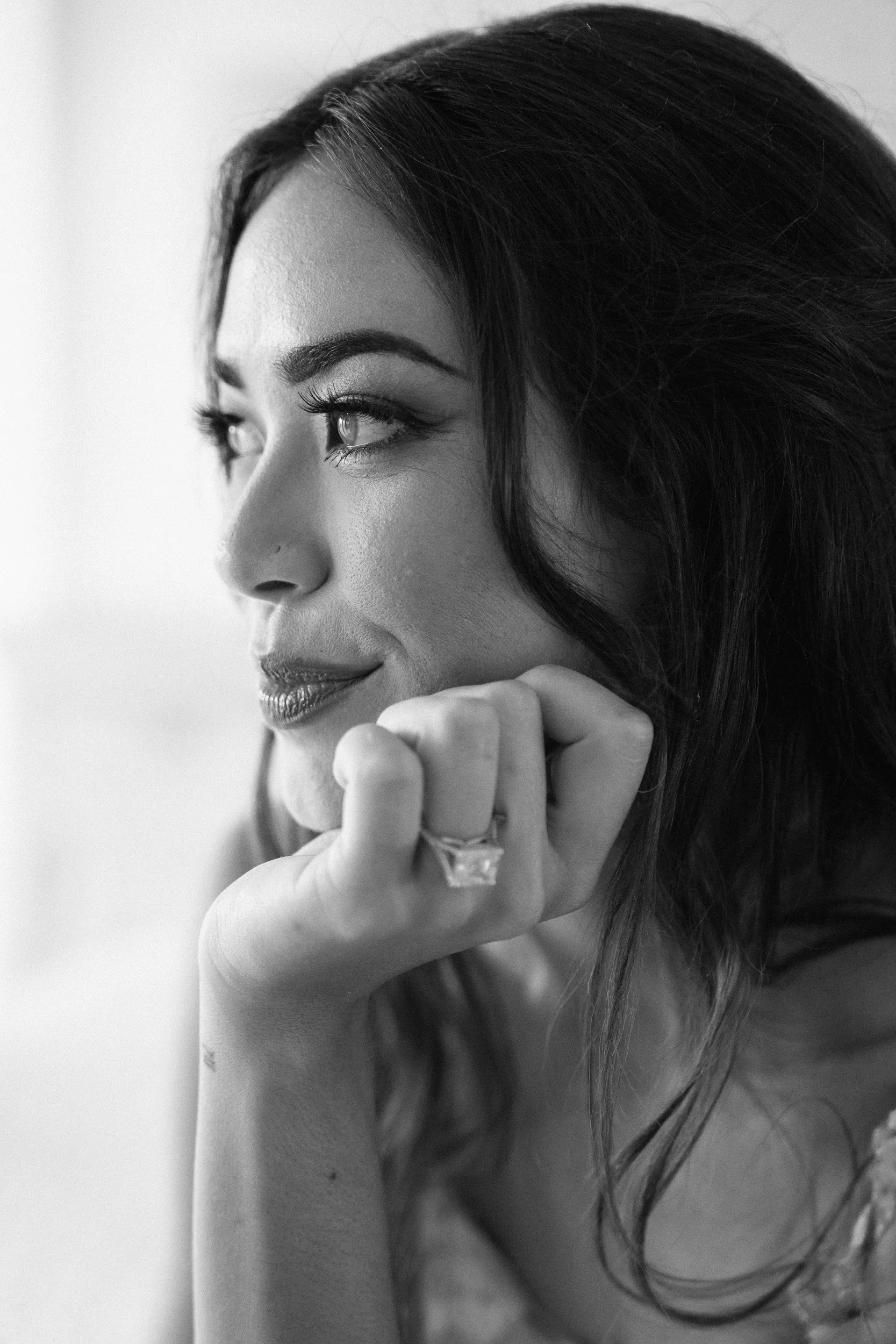 Black and white photo of a woman with long dark hair, looking to her left with her hand resting near her chin, wearing a ring.