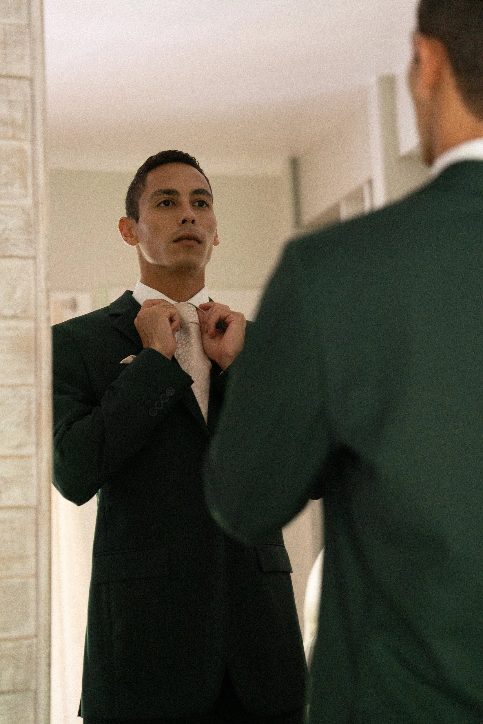 A man adjusting his tie in front of a mirror, dressed in a black suit and white shirt, with another person visible partially in the foreground.