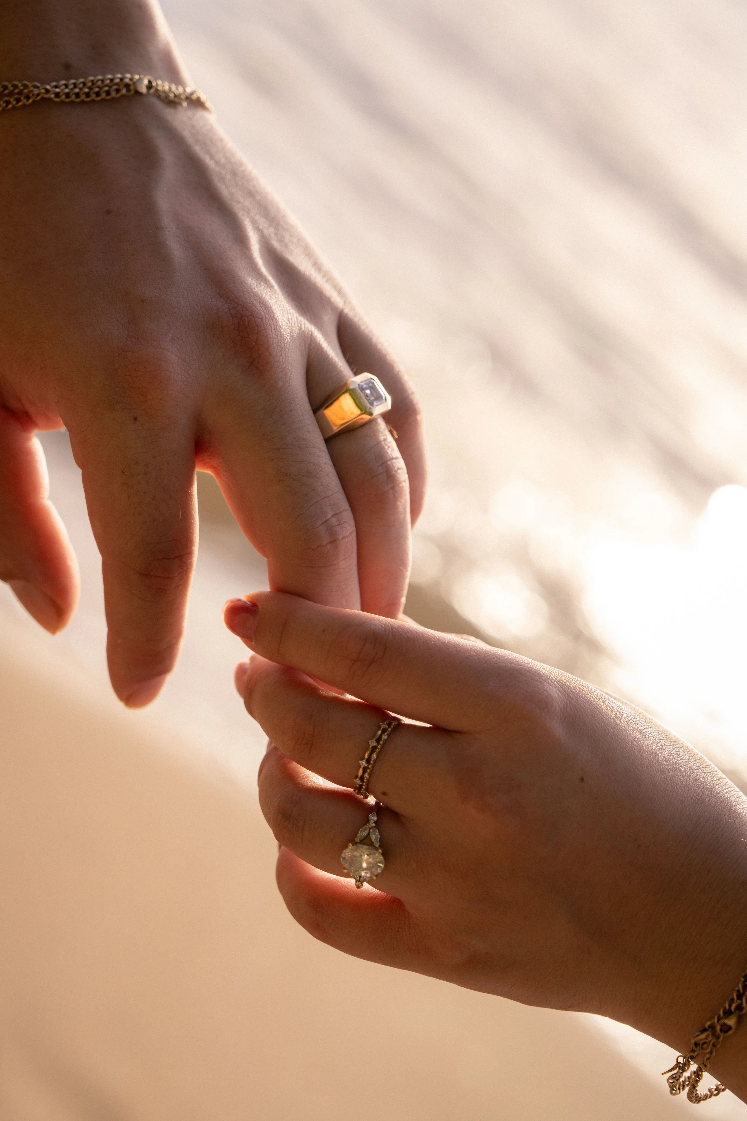 Close-up of two hands with jewelry, including a ring with a large clear stone, a gold chain bracelet, and a delicate ring with a small stone, against a soft background.