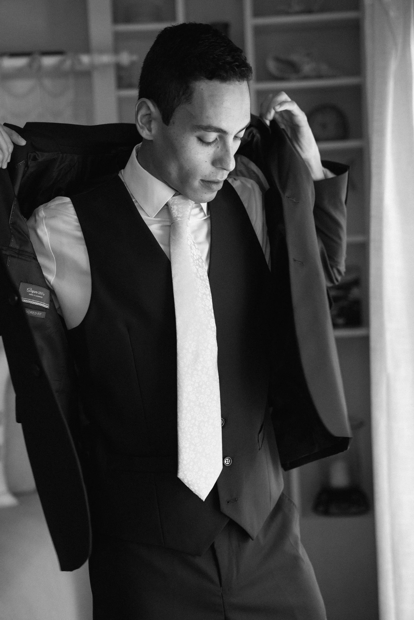 Black and white photo of a young man putting on a suit jacket, dressed in a white shirt, light-colored tie, and dark pants, standing in a room with shelves in the background.