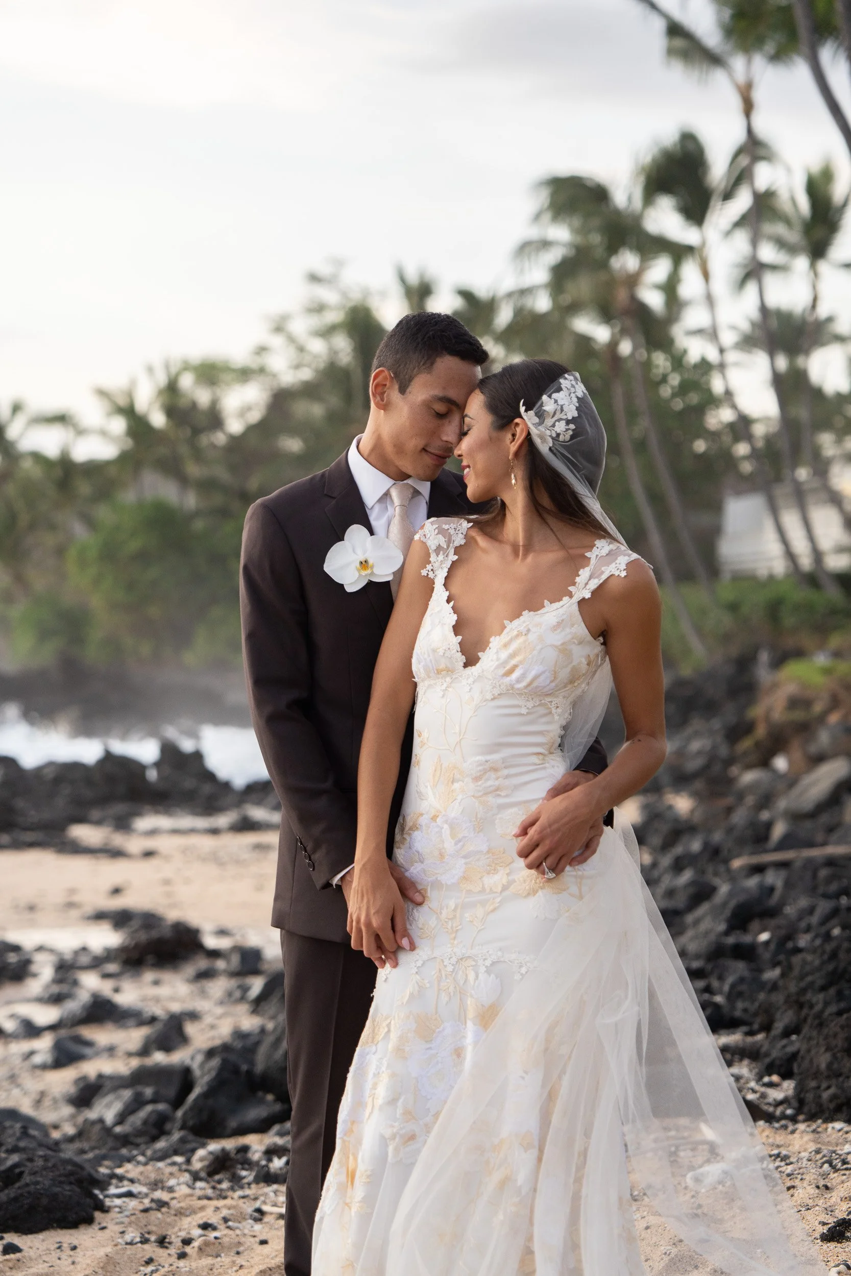 A bride and groom on a rocky beach, standing close with foreheads touching, dressed in wedding attire. The bride wears a white lace wedding gown and veil with floral details, while the groom wears a dark suit with a white shirt and tie, sporting a la