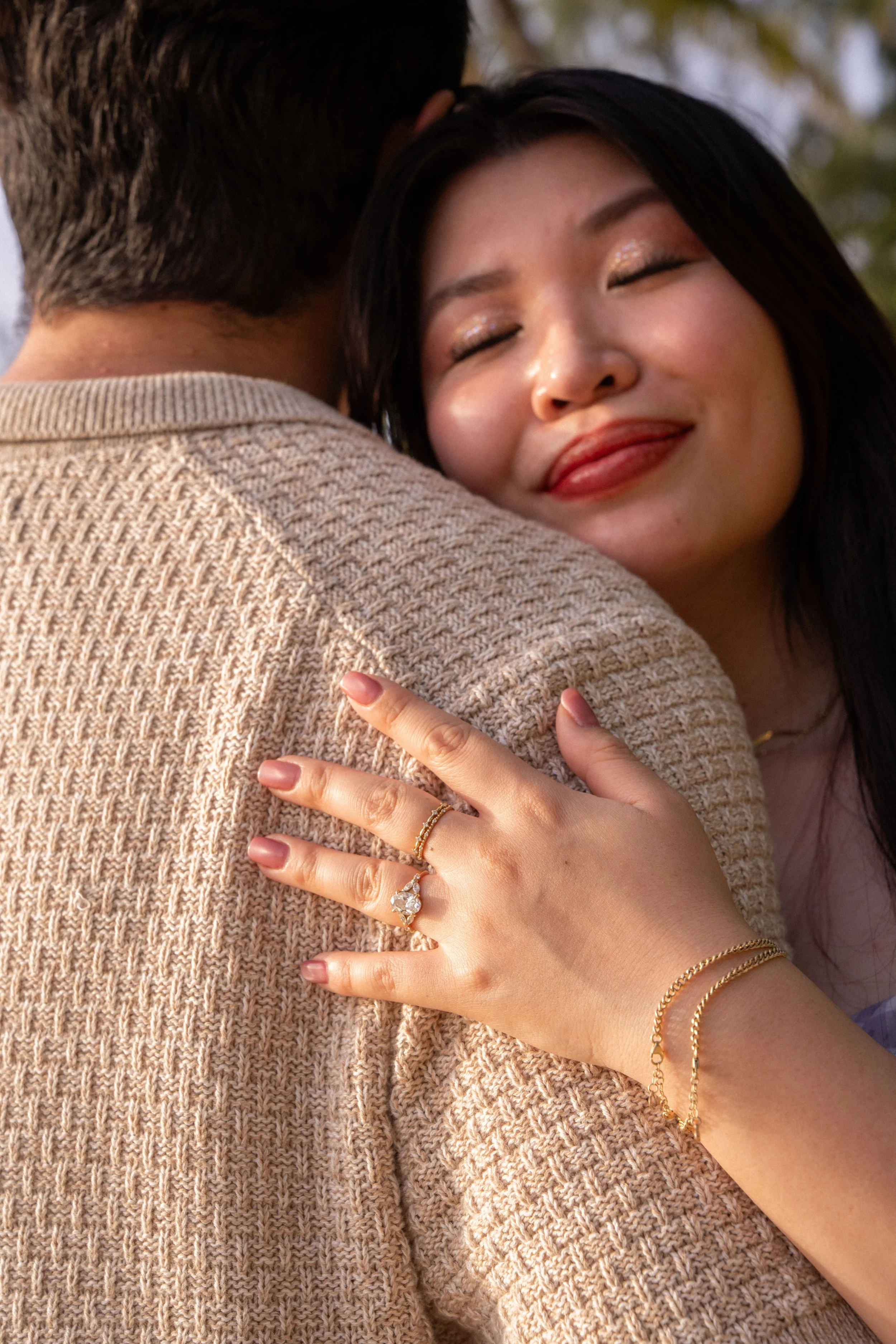 Close-up of a woman with dark hair smiling with her eyes closed, wearing makeup, and embracing someone wearing a beige knitted sweater. The woman's hand, which is resting on the sweater, is adorned with rings and gold bracelets.