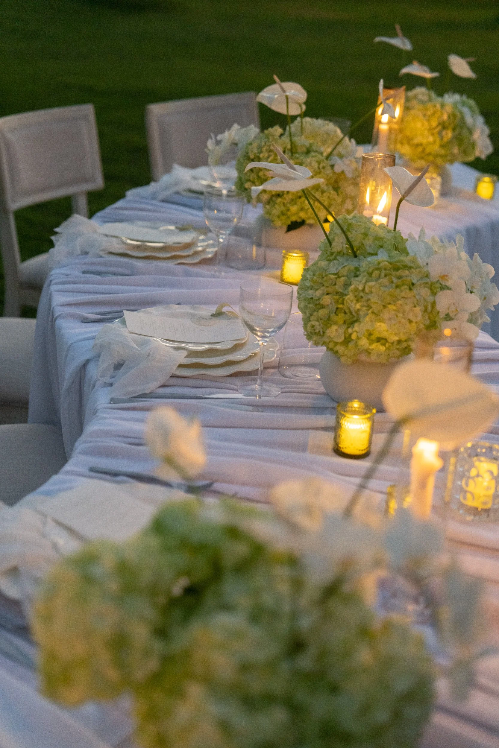 A long white table decorated with white floral centerpieces and candles, set for an outdoor evening event with chairs and tableware.