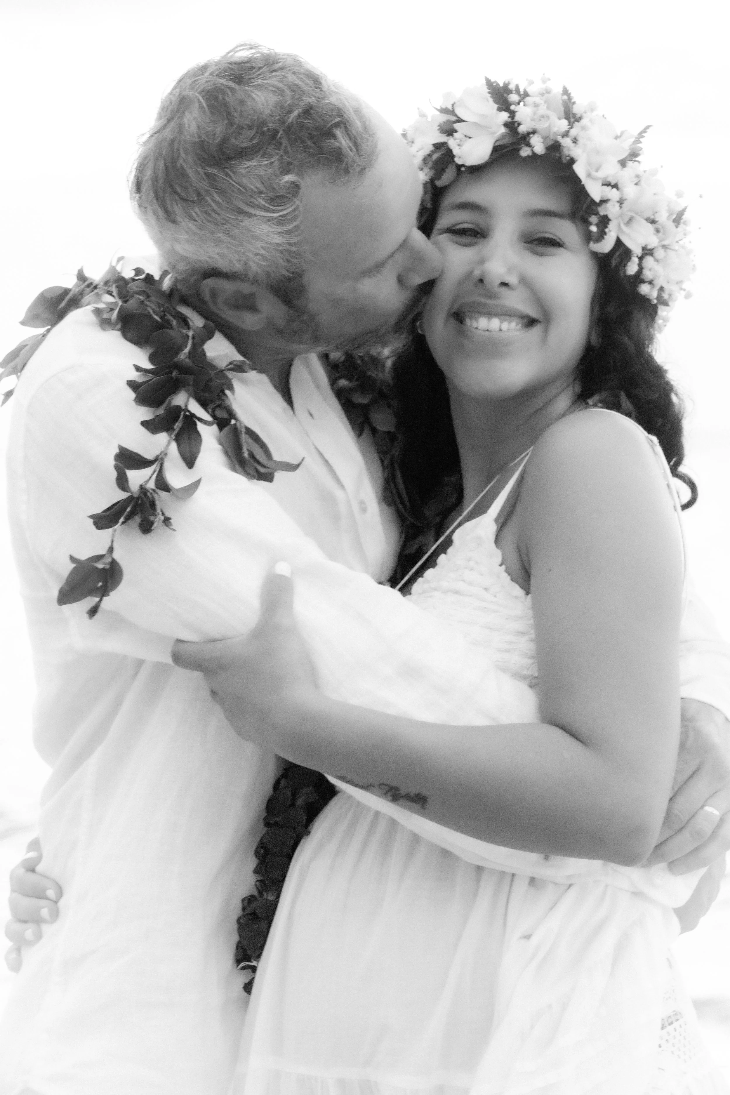 A couple on their wedding day, with the man giving a kiss on the woman's cheek. The woman is smiling, wearing a flower crown and a white dress, while the man is in a white shirt with a floral lei around his neck.