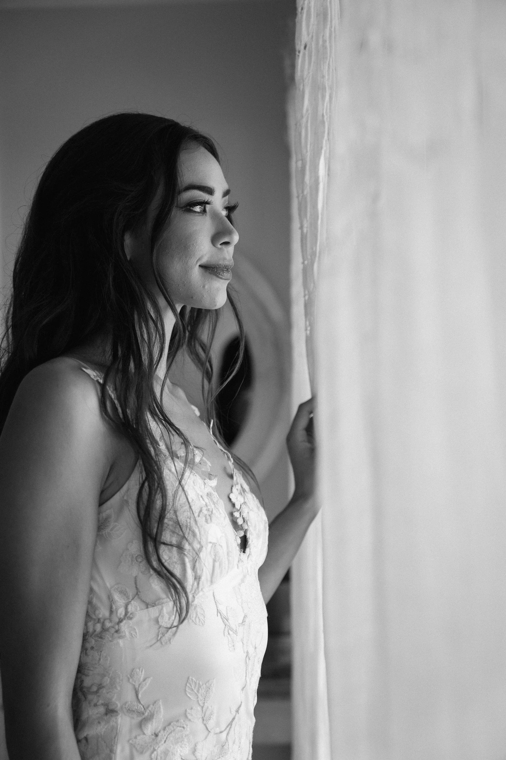 A woman with long, wavy hair looking out a window. She has a slight smile and is wearing a sleeveless, lace dress with floral embroidery.