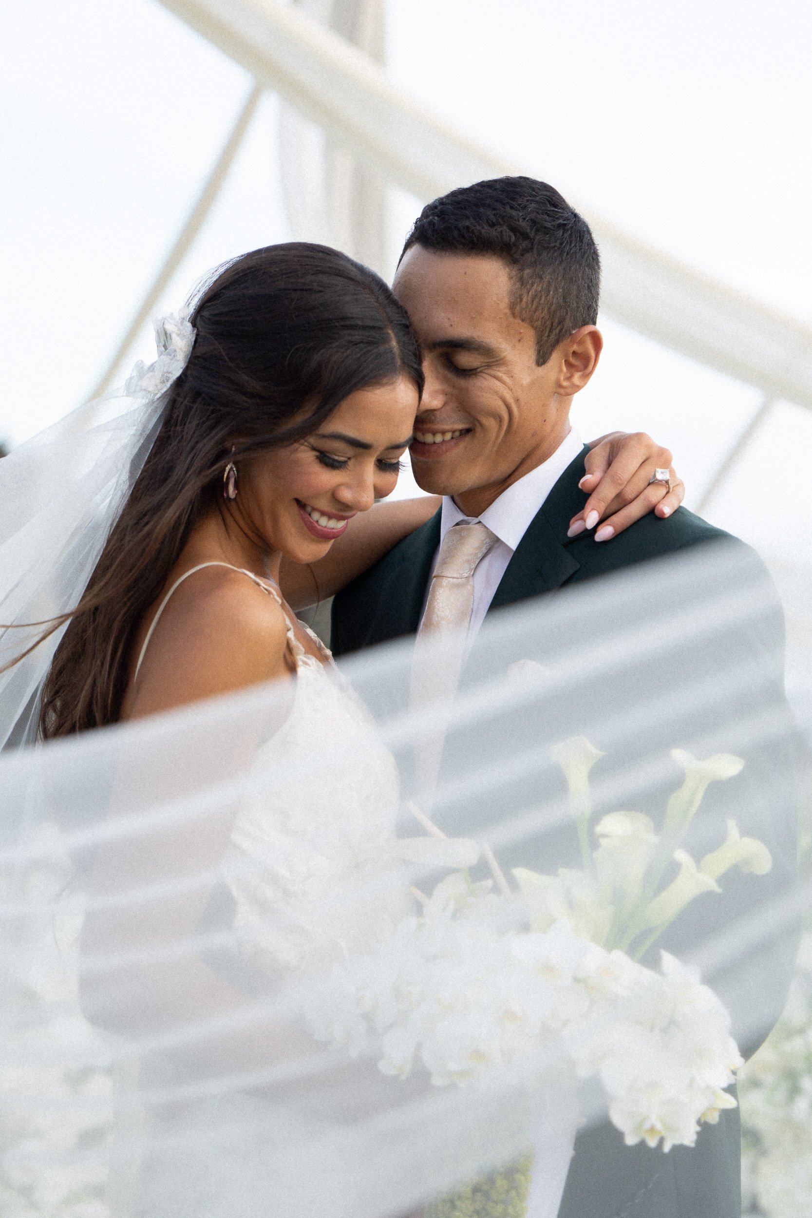 A close-up of a bride and groom smiling and holding each other during their wedding, with the bride wearing a veil and jewelry, and the groom dressed in a suit and tie.
