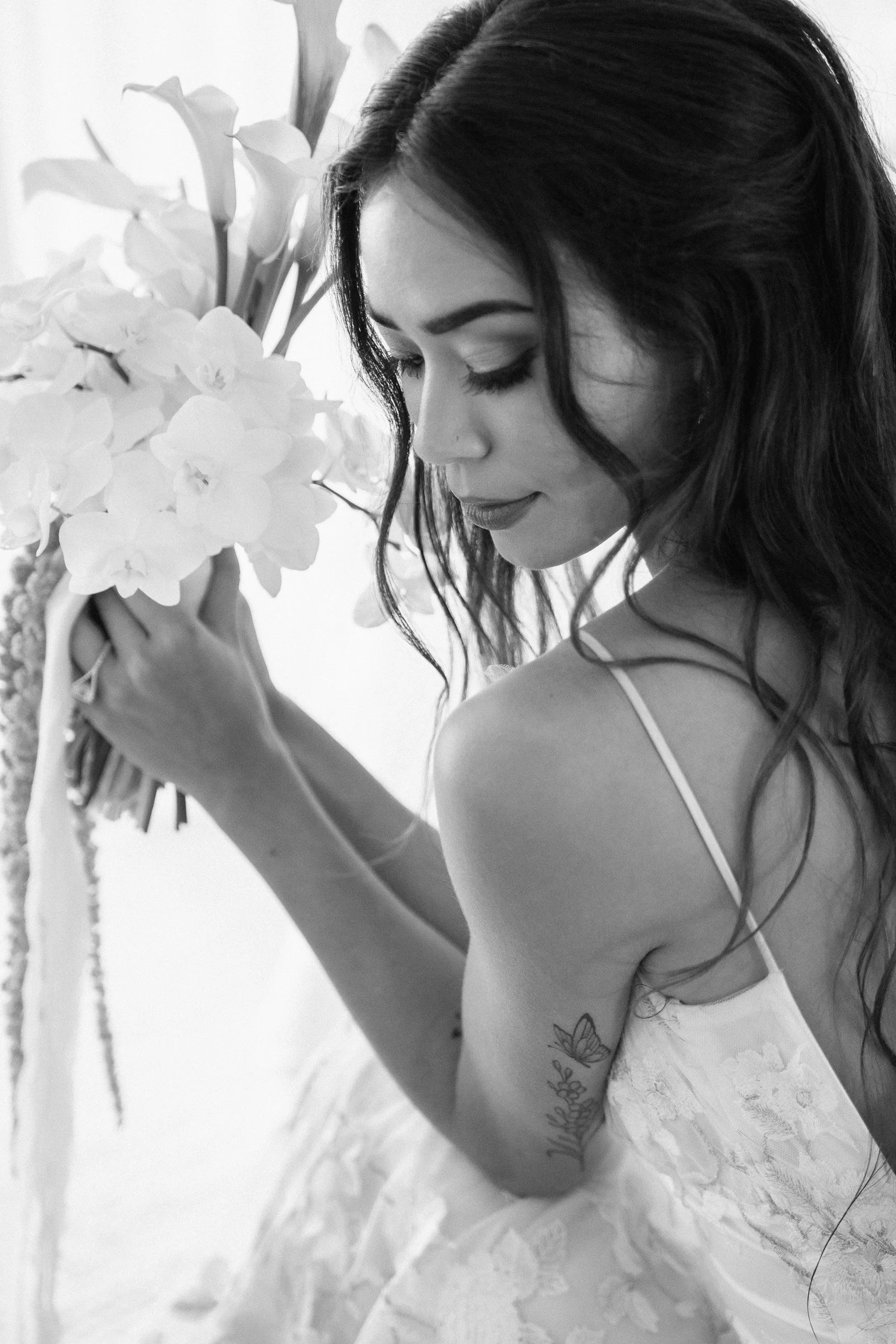 Black and white photo of a woman with long, wavy hair and makeup, holding a bouquet of white flowers, wearing a white dress with thin straps, and showing a tattoo on her arm.
