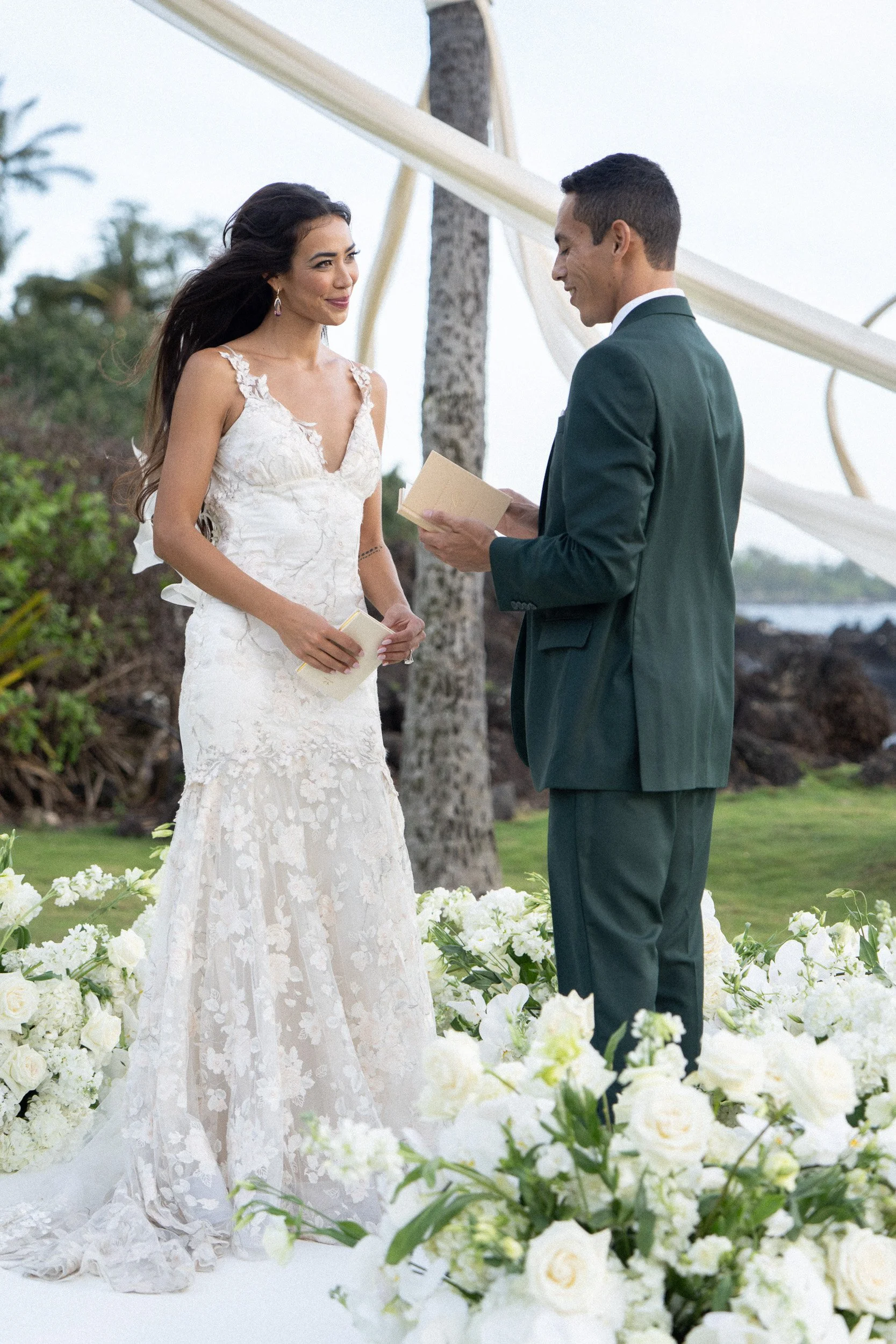 Couple exchanging vows during outdoor wedding ceremony with floral decorations and palm trees in the background.