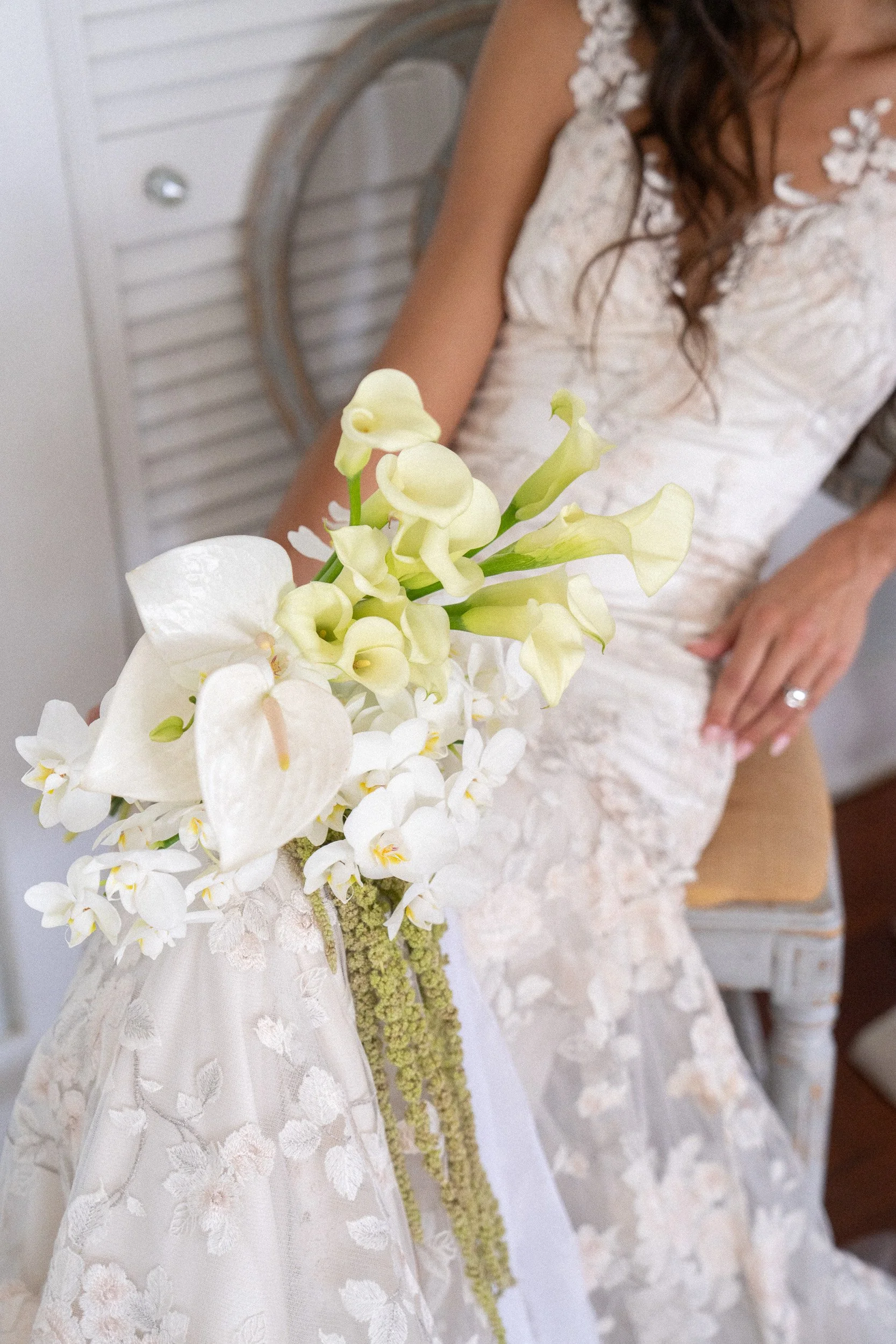 Close-up of a woman in a white lace wedding dress holding a bouquet of white and cream flowers, including orchids and calla lilies, with a woman in a white dress sitting in the background.