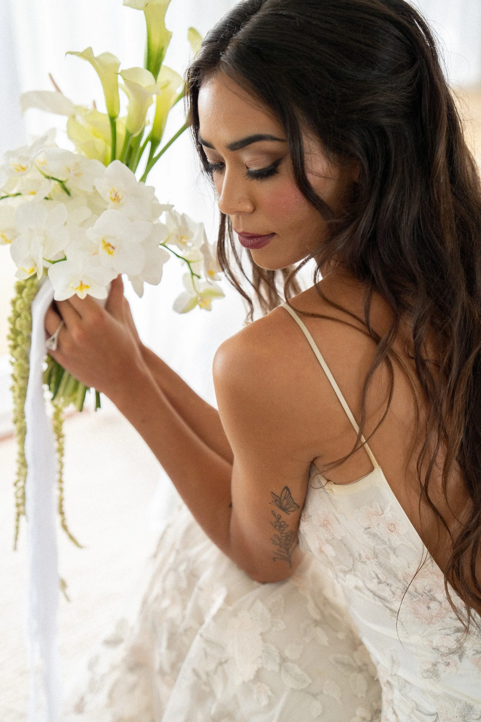 A woman with dark hair wearing a white lace dress, holding a bouquet of white flowers, and looking down.