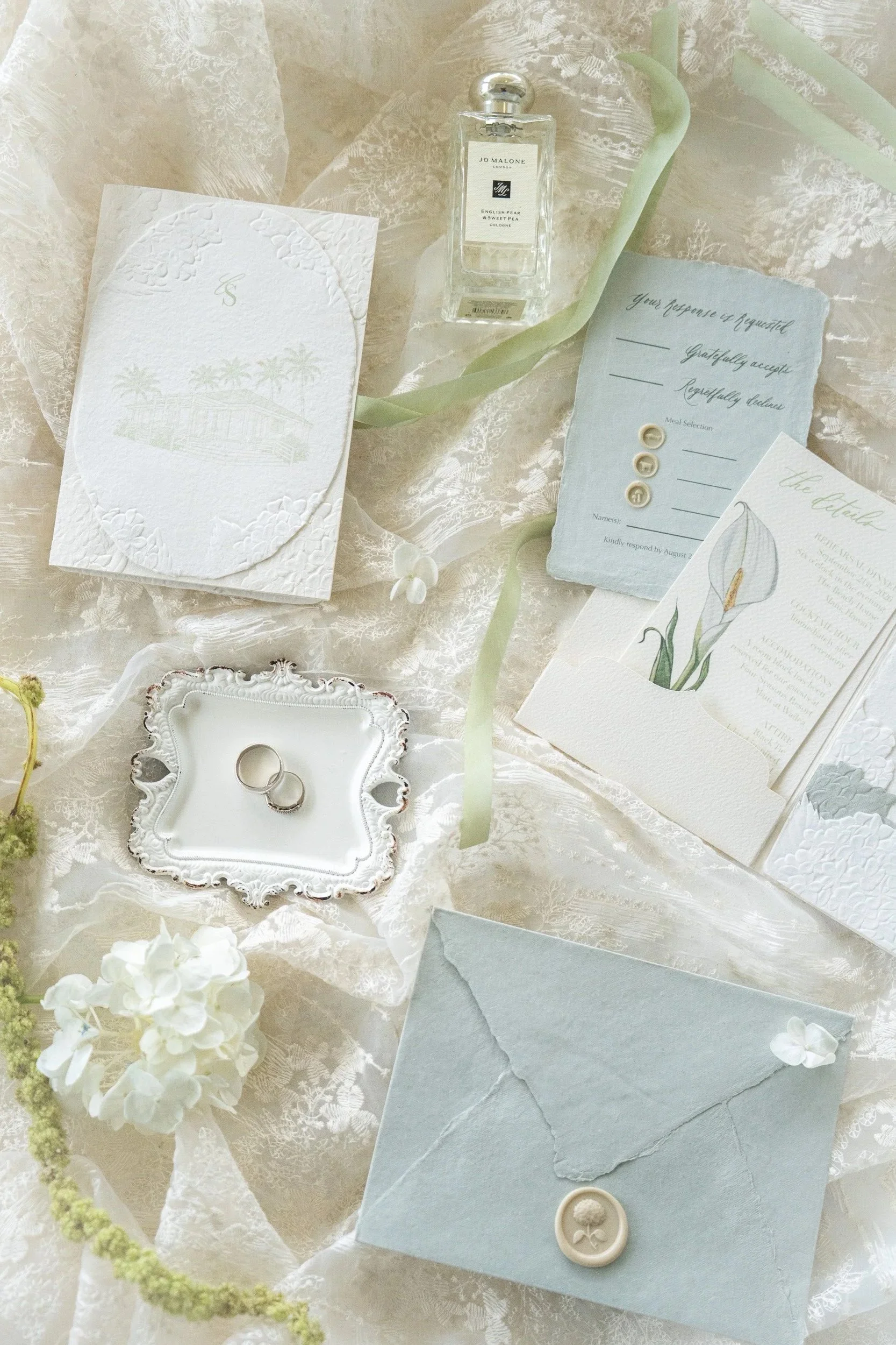 A wedding invitation suite on a lace tablecloth includes an envelope, a small card with a floral illustration, a telegram with options for response, a perfume bottle labeled 'Jo Malone English Pear & Sweet Pea,' a small ornate tray with wedding rings