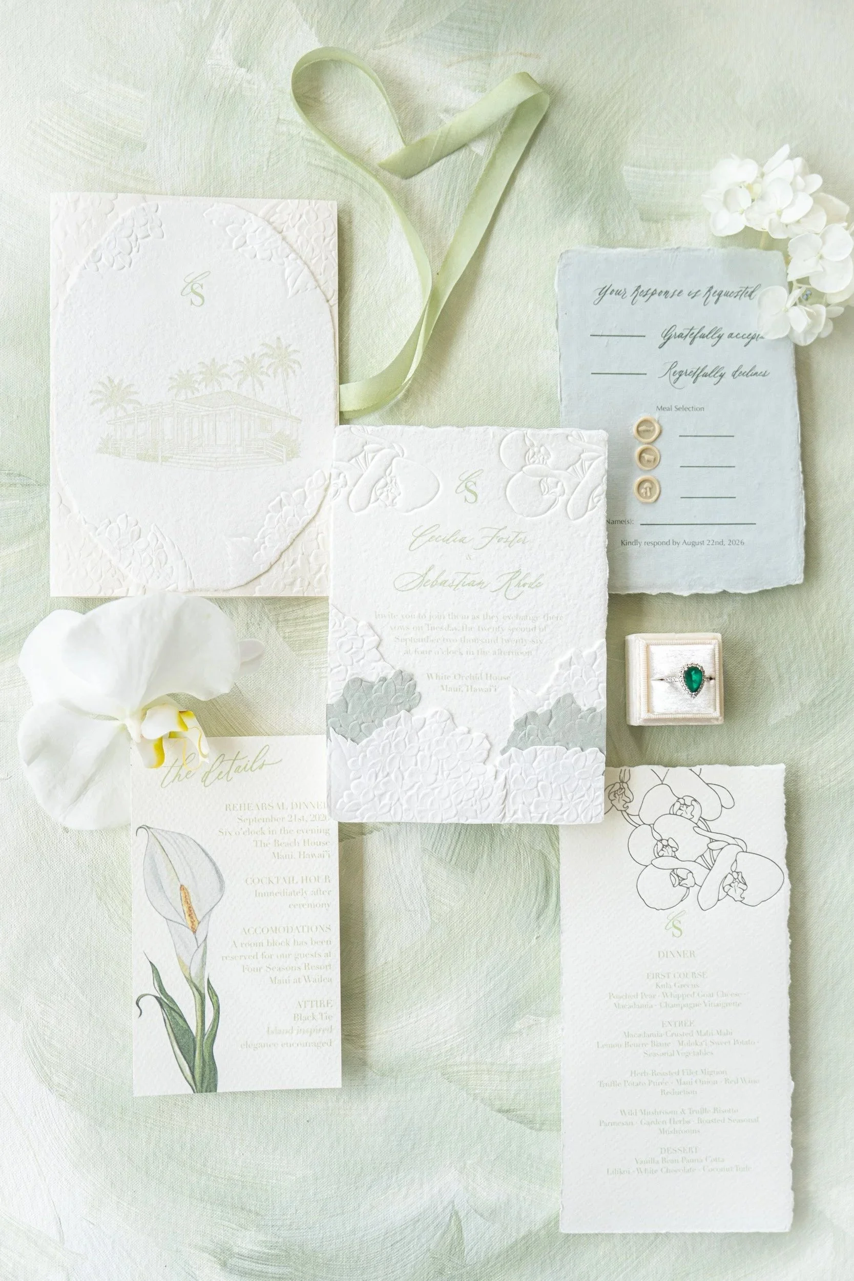 A flat-lay of wedding stationery items, including invitation cards, RSVP card, menu, and a ring box with a green gemstone. The setup features a light green background with floral accents and white orchids.