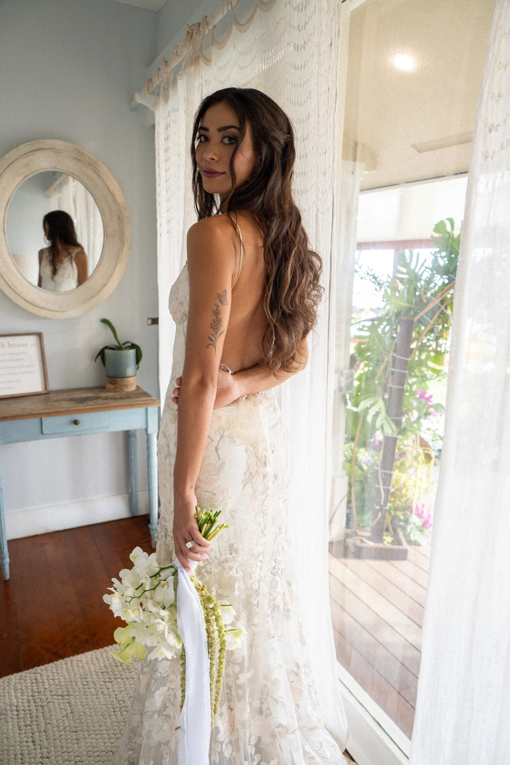 A woman with long wavy hair, wearing a white lace dress, stands indoors near a window with sheer curtains, holding a bouquet of white flowers.