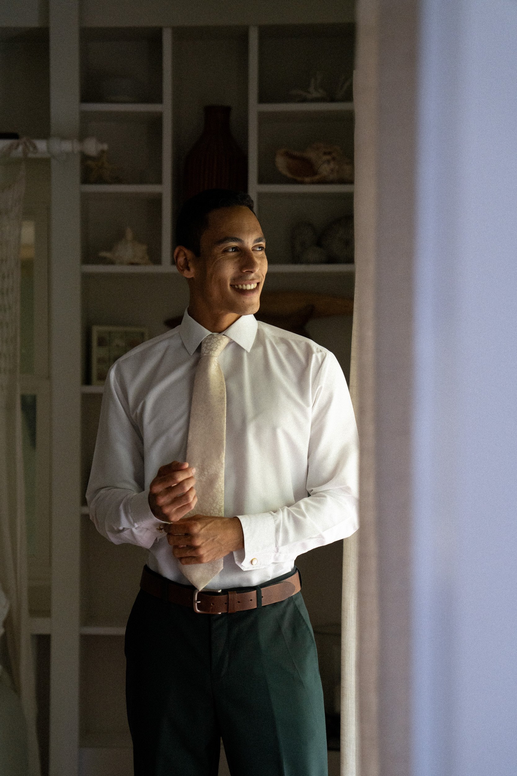 A man smiling while adjusting his beige tie, wearing a white dress shirt and dark pants, standing near a window with a curtain in a room with a bookshelf containing decorative items.