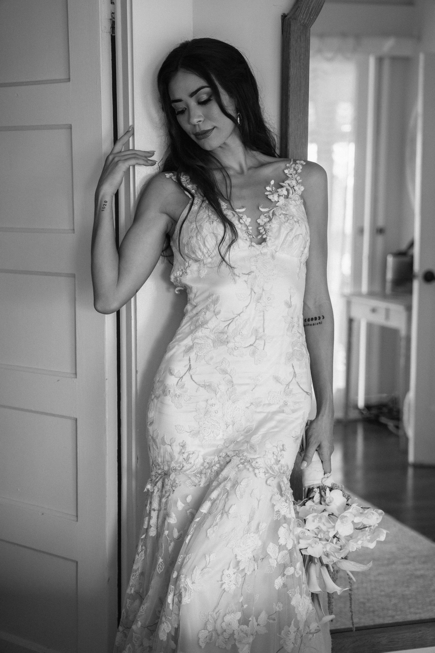 Black and white photo of a woman in an elegant lace wedding dress, holding a bouquet of flowers, standing near a door and a mirror, with a serene expression.