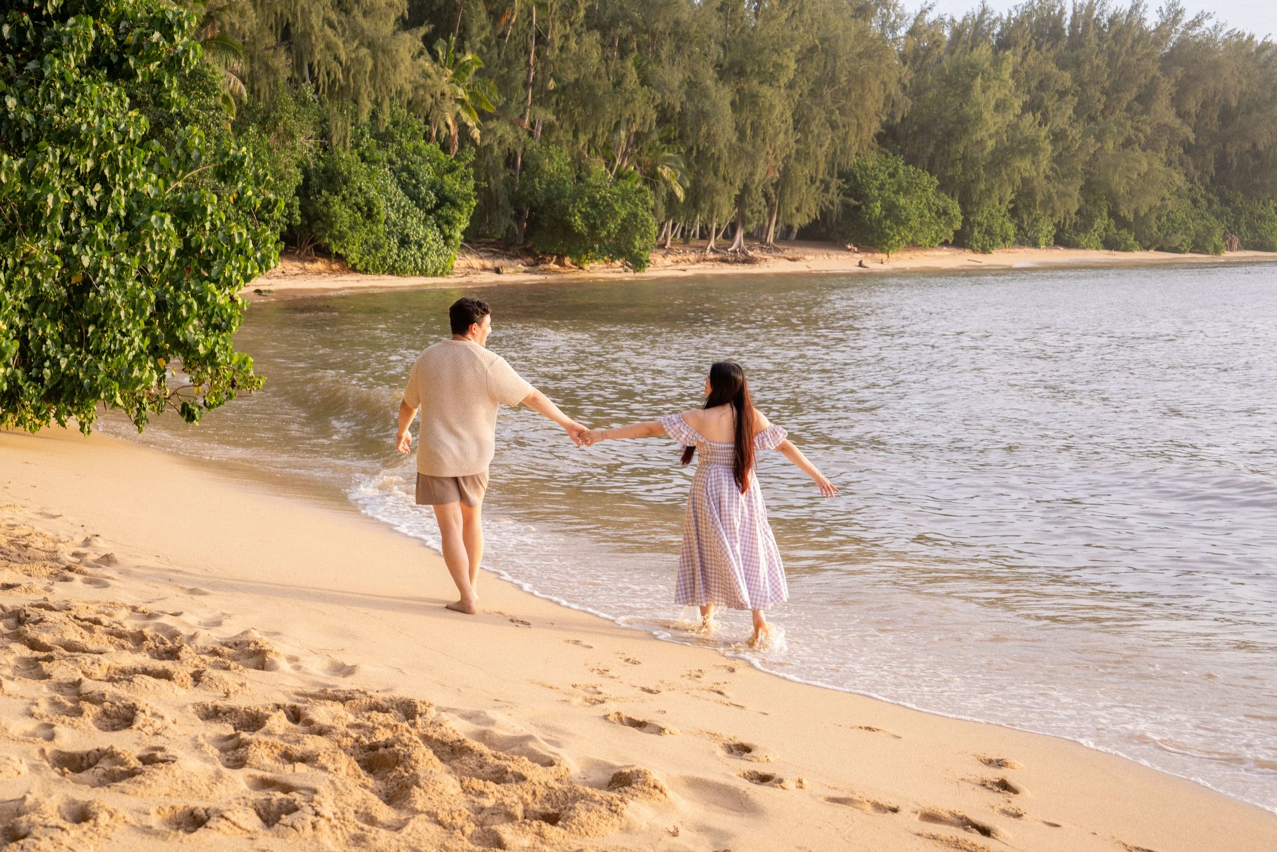 A man and a woman holding hands and walking into the water at the beach during sunset, with lush green trees in the background.