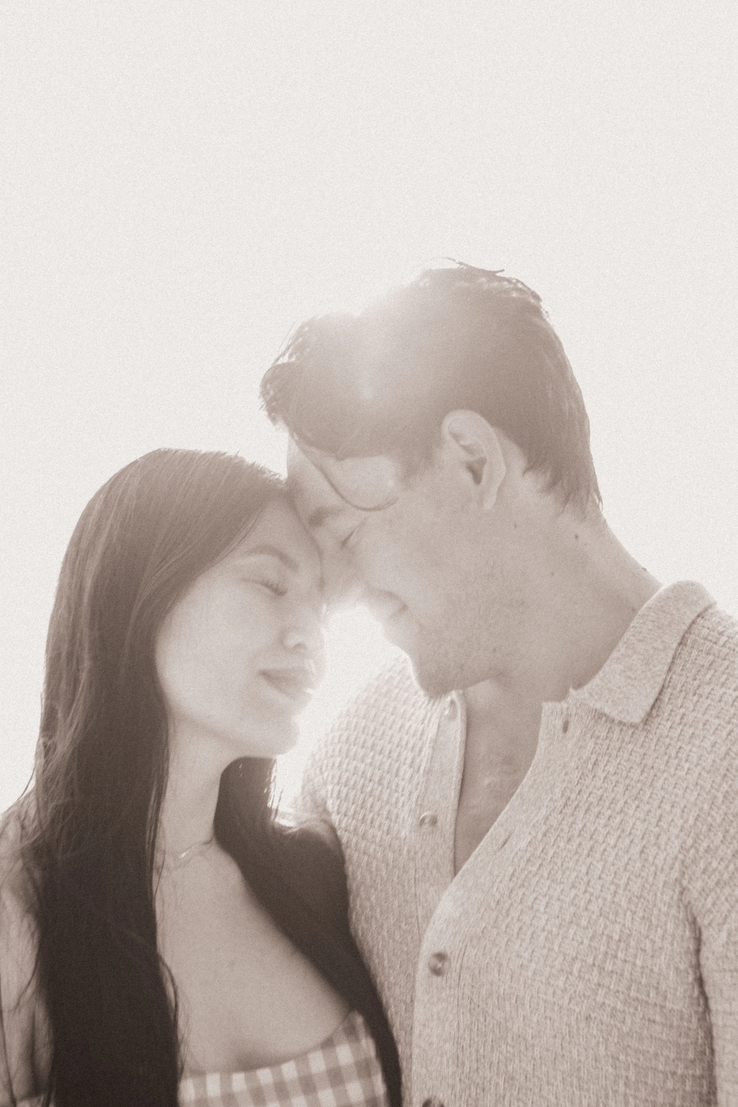 A close-up photo of a man and woman with their foreheads touching, eyes closed, smiling gently, with soft backlighting creating a romantic mood.