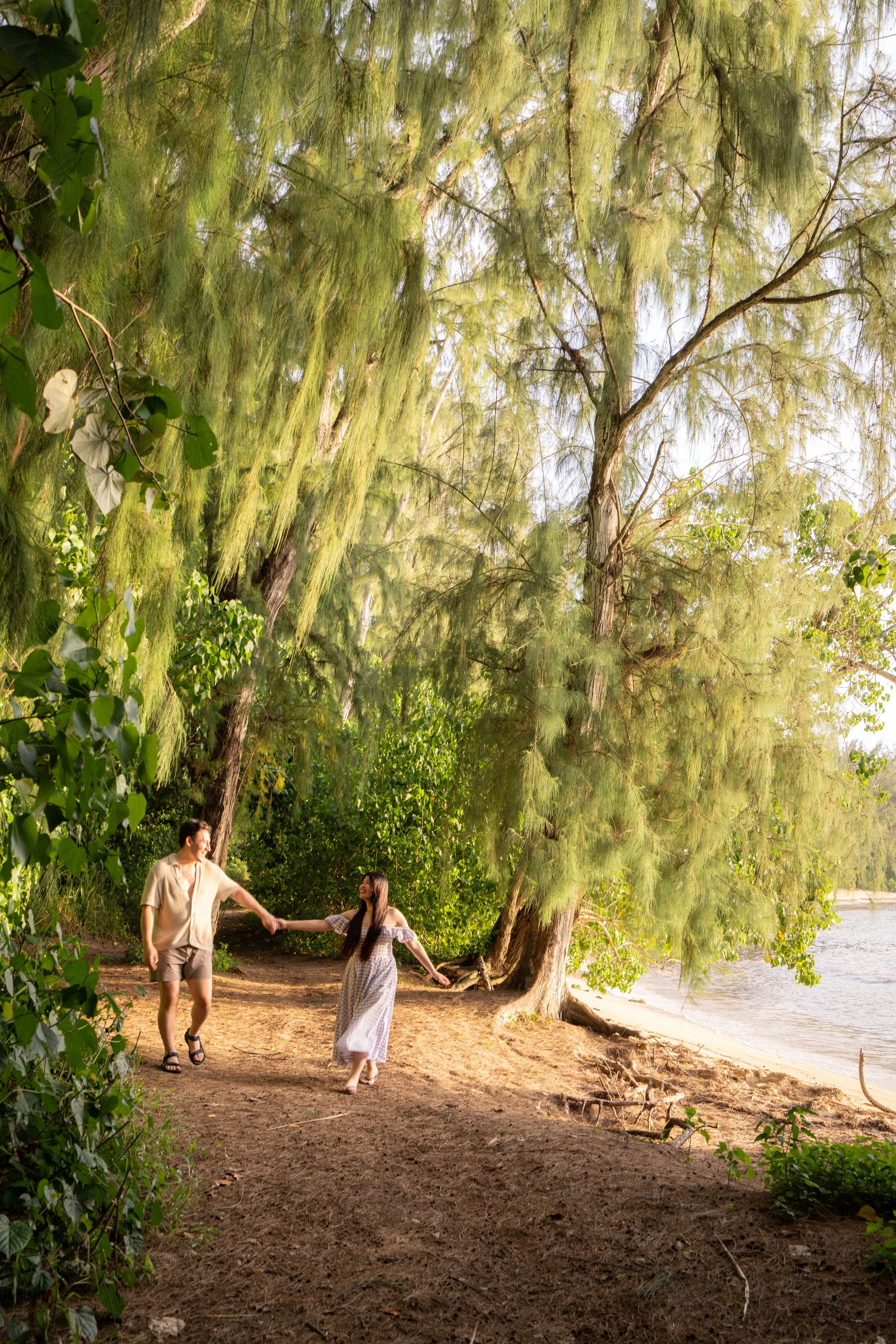 A young couple holding hands and walking on a sandy beach path surrounded by tall trees with green foliage near the water.