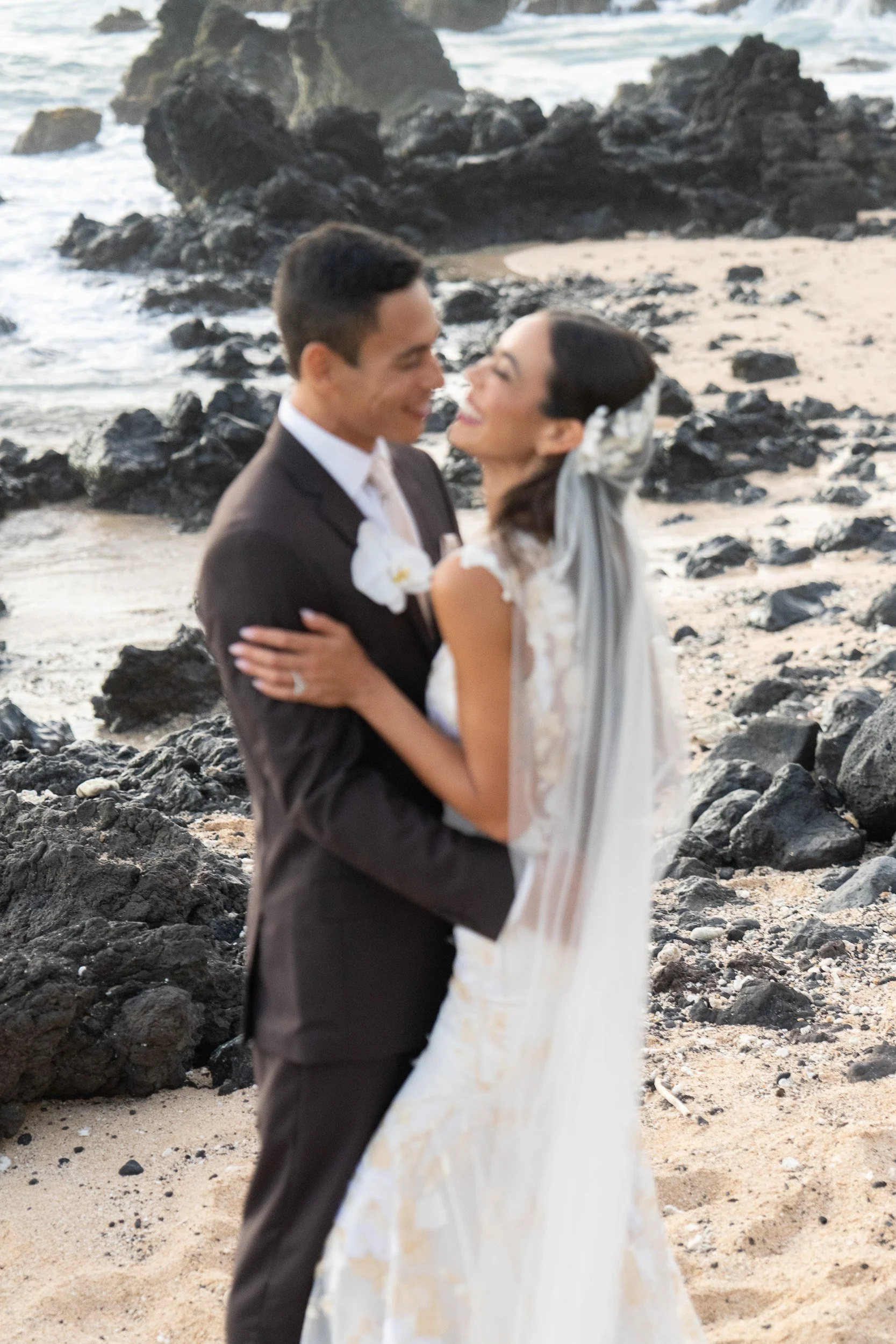 A couple in wedding attire standing on a rocky beach, gazing at each other happily.