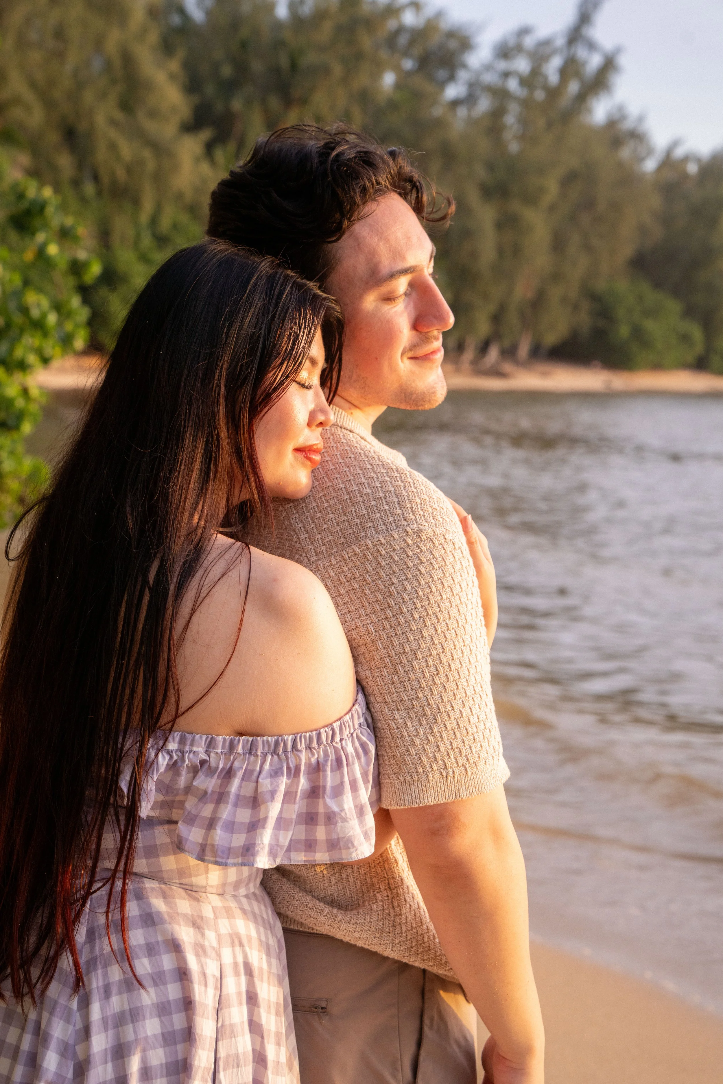 A man and a woman stand close together near the water's edge with their eyes closed, enjoying the peaceful moment at sunset or sunrise by a river or lake. The woman has long dark hair and is wearing a pinkish checkered off-shoulder dress, while the m