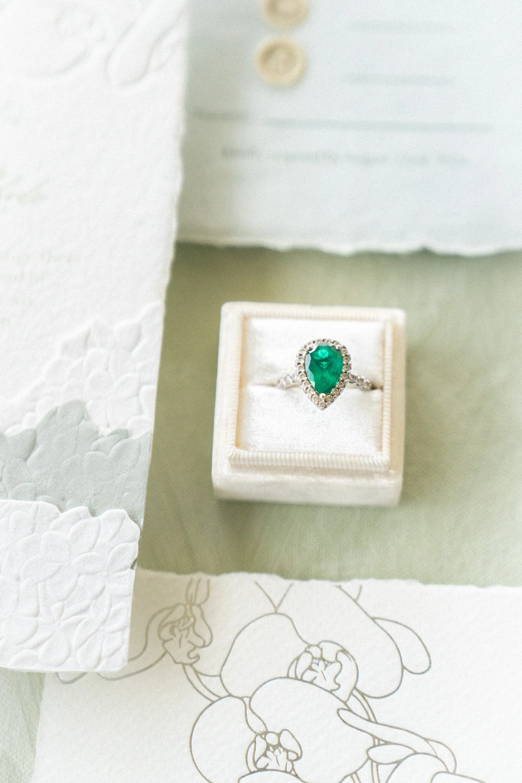Close-up of a ring with a pear-shaped green gemstone surrounded by small diamonds, placed in a white jewelry box.