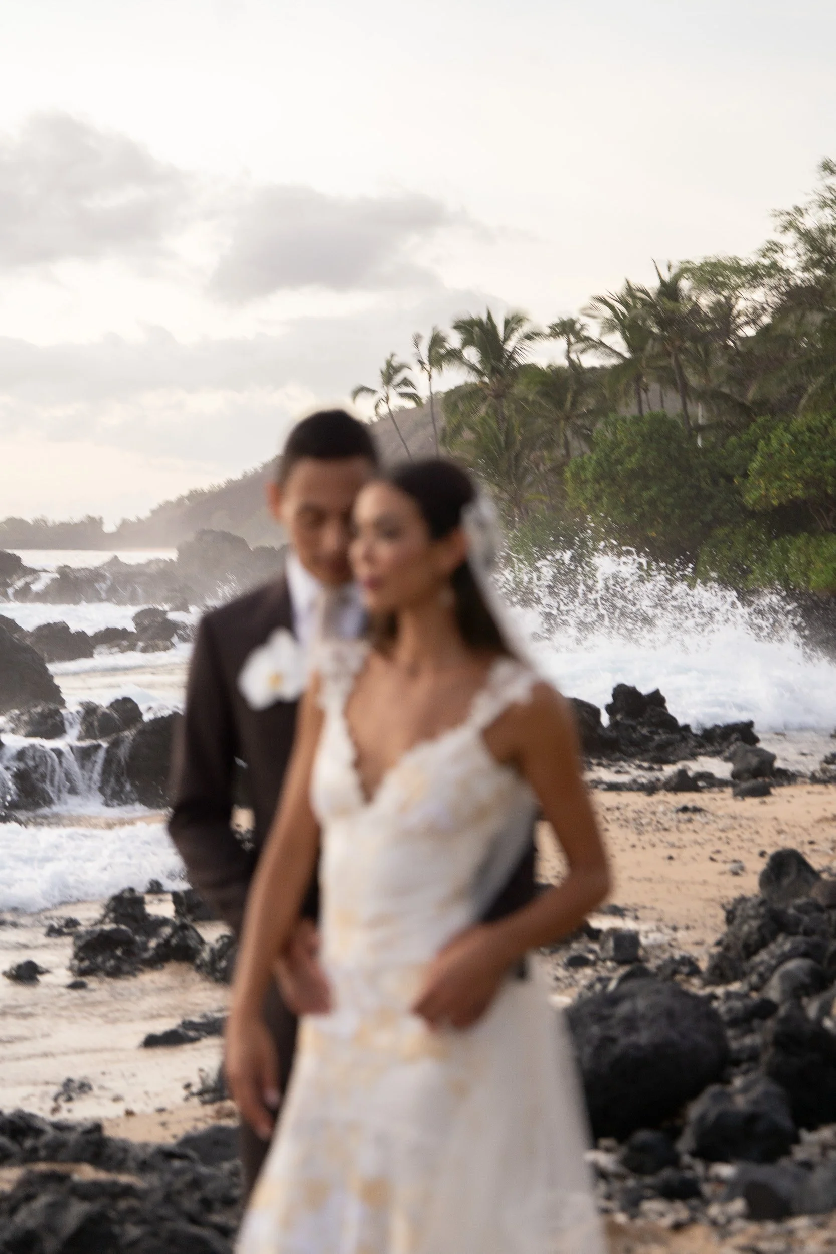 A blurred photo of a couple in wedding attire standing on a rocky beach with palm trees and ocean waves in the background.