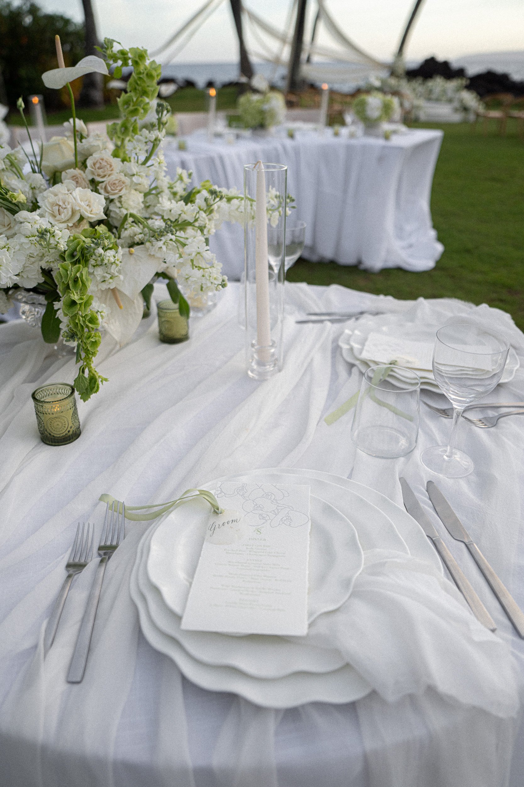 A beautifully set wedding reception table with white tablecloth, glassware, silverware, and a floral centerpiece of white and green flowers. There are candles and a menu card on a plate, and a backdrop of white tent with a scenic outdoor landscape.