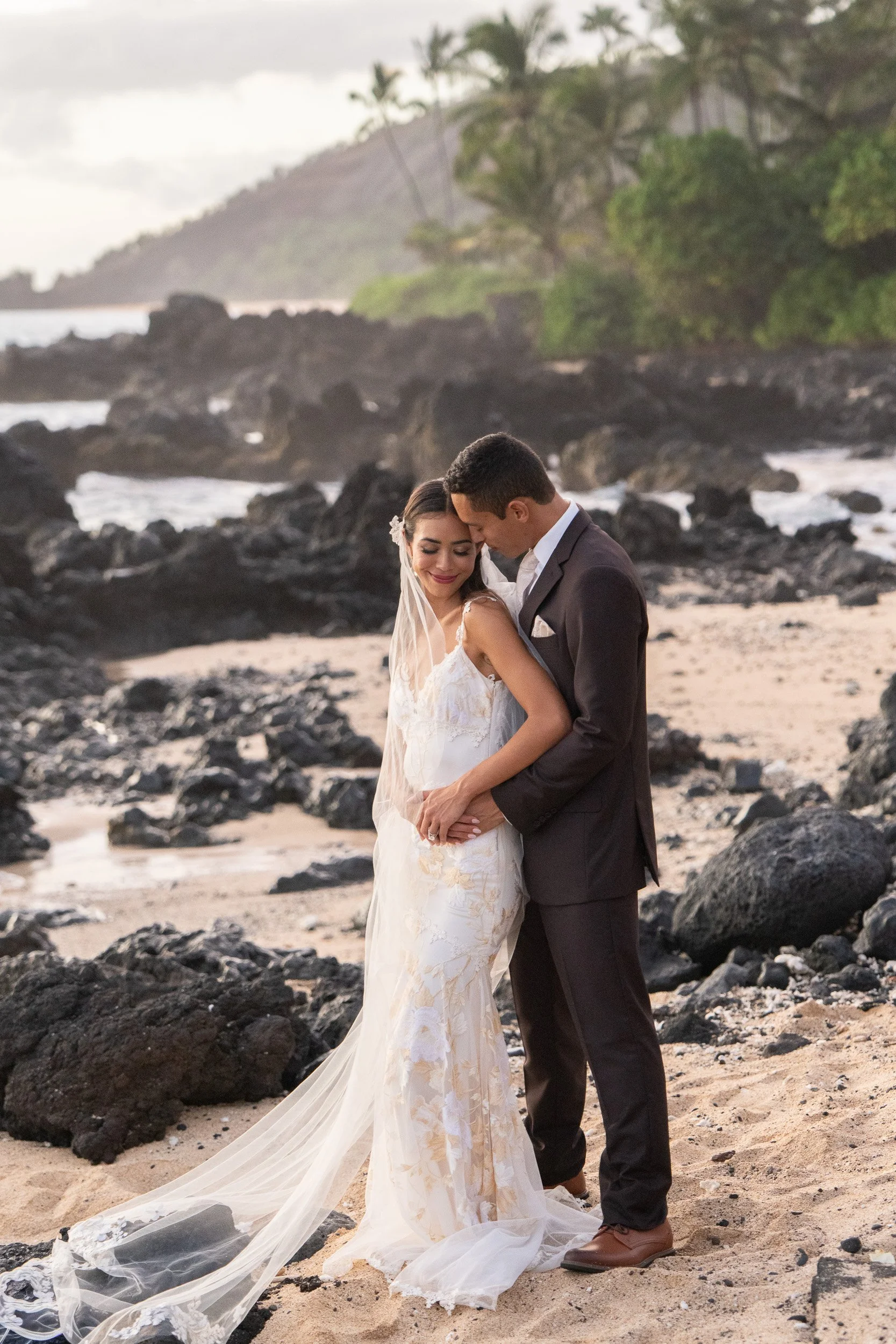 A newlywed couple sharing an intimate moment on a rocky beach at sunset, with lush greenery and distant mountains in the background.