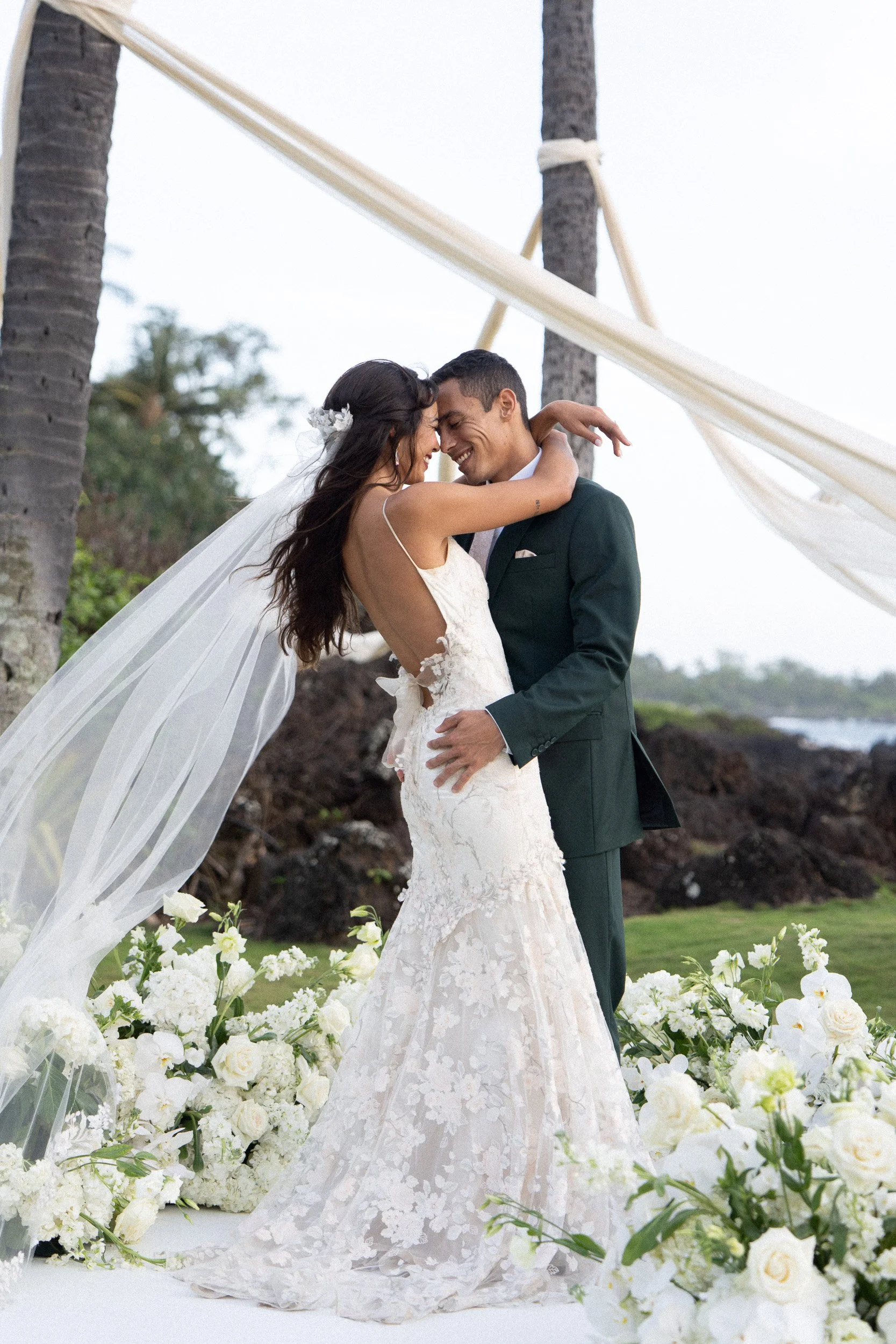 A bride and groom embrace on a beach wedding altar decorated with white flowers and greenery, with tall palm trees and a cloudy sky in the background.