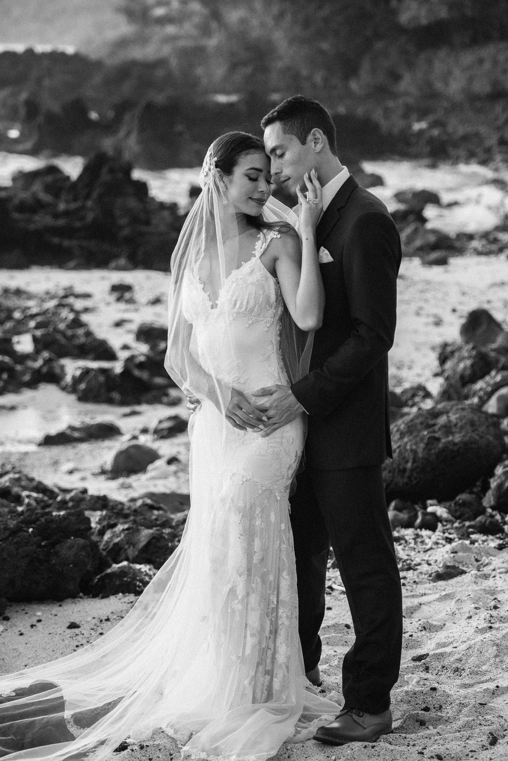 A black-and-white photograph of a bride and groom standing close together on a rocky beach, with the bride in a wedding dress and veil, and the groom in a suit, both with their eyes closed, sharing an intimate moment.