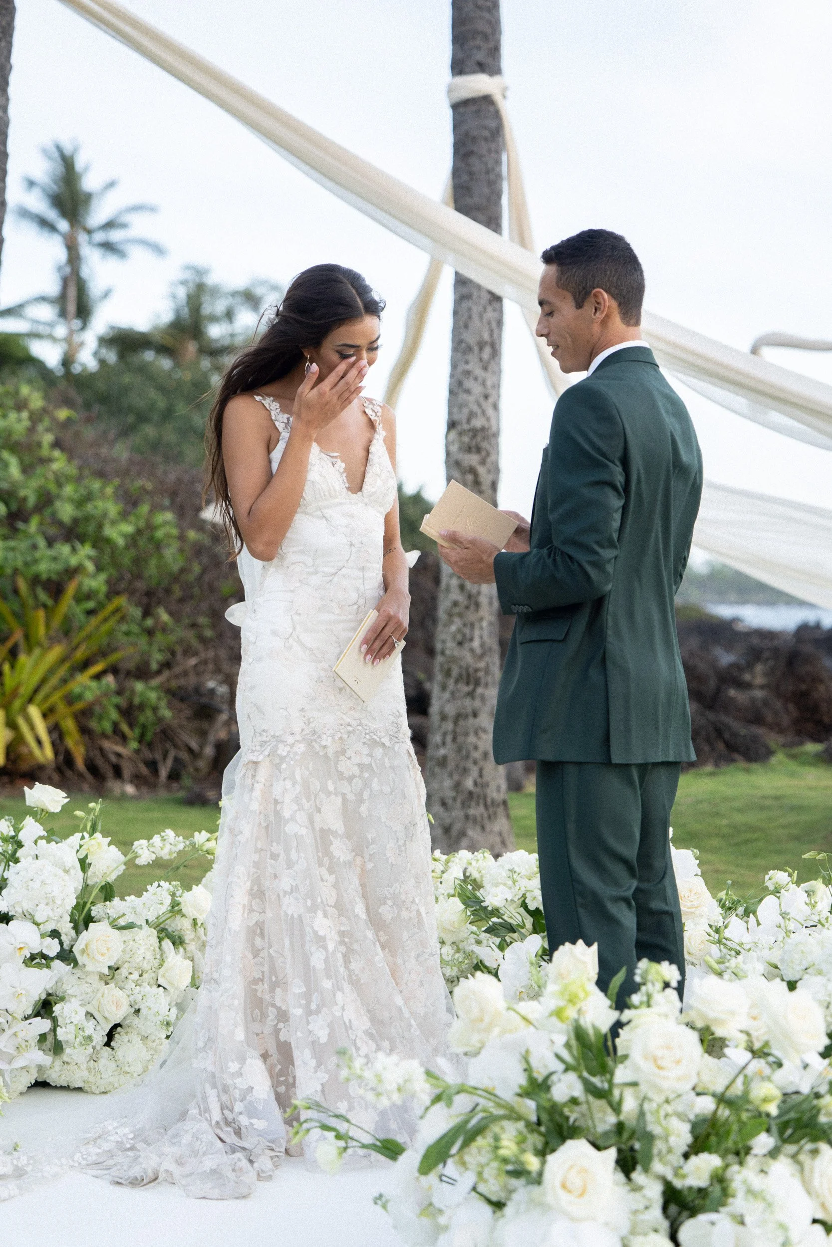 A bride and groom stand together during an outdoor wedding ceremony surrounded by white flowers, with the bride covering her mouth and tearfully smiling while holding a book, and the groom holding a ring box and smiling.