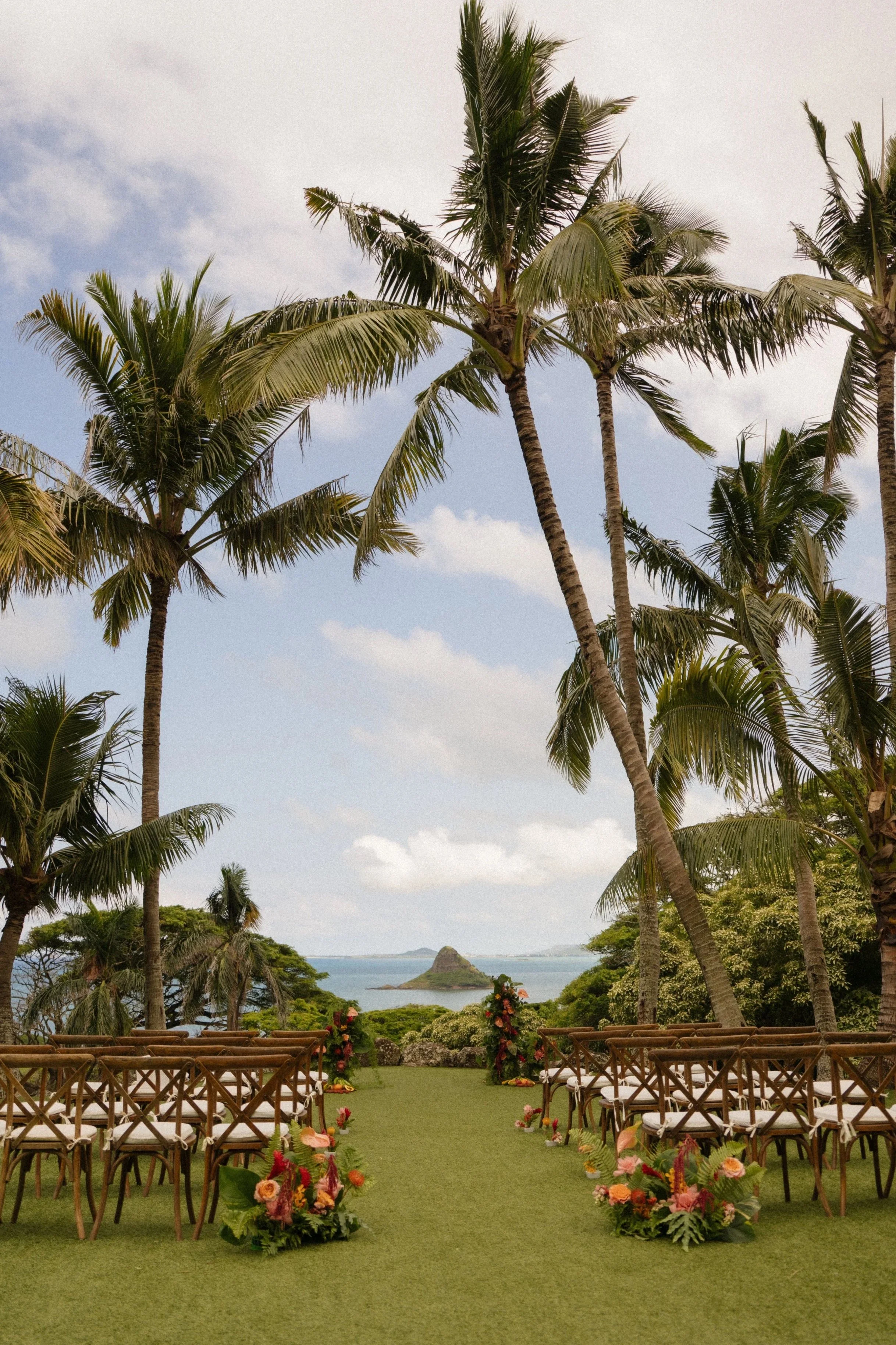 Outdoor wedding setup with rows of chairs on a green aisle leading to an altar decorated with flowers, overlooking the ocean with palm trees and a distant island in the background.
