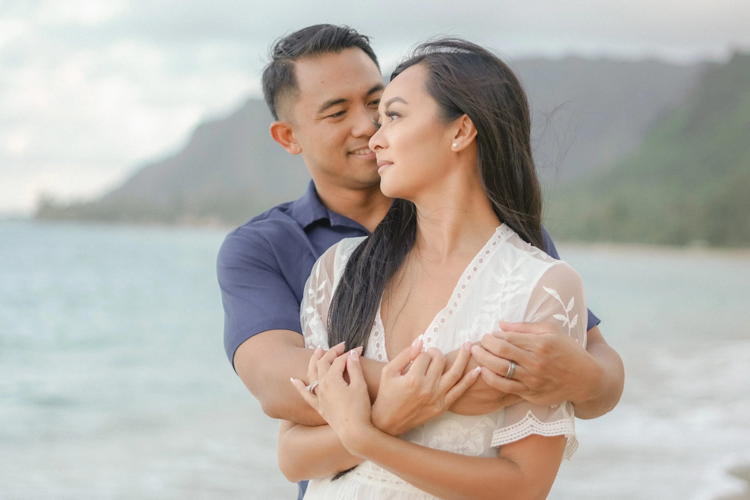A couple embracing on a beach, with mountains and water in the background, close-up of their faces and hands. intimate elopement ceremony in Hawaii captured by isis monteux
