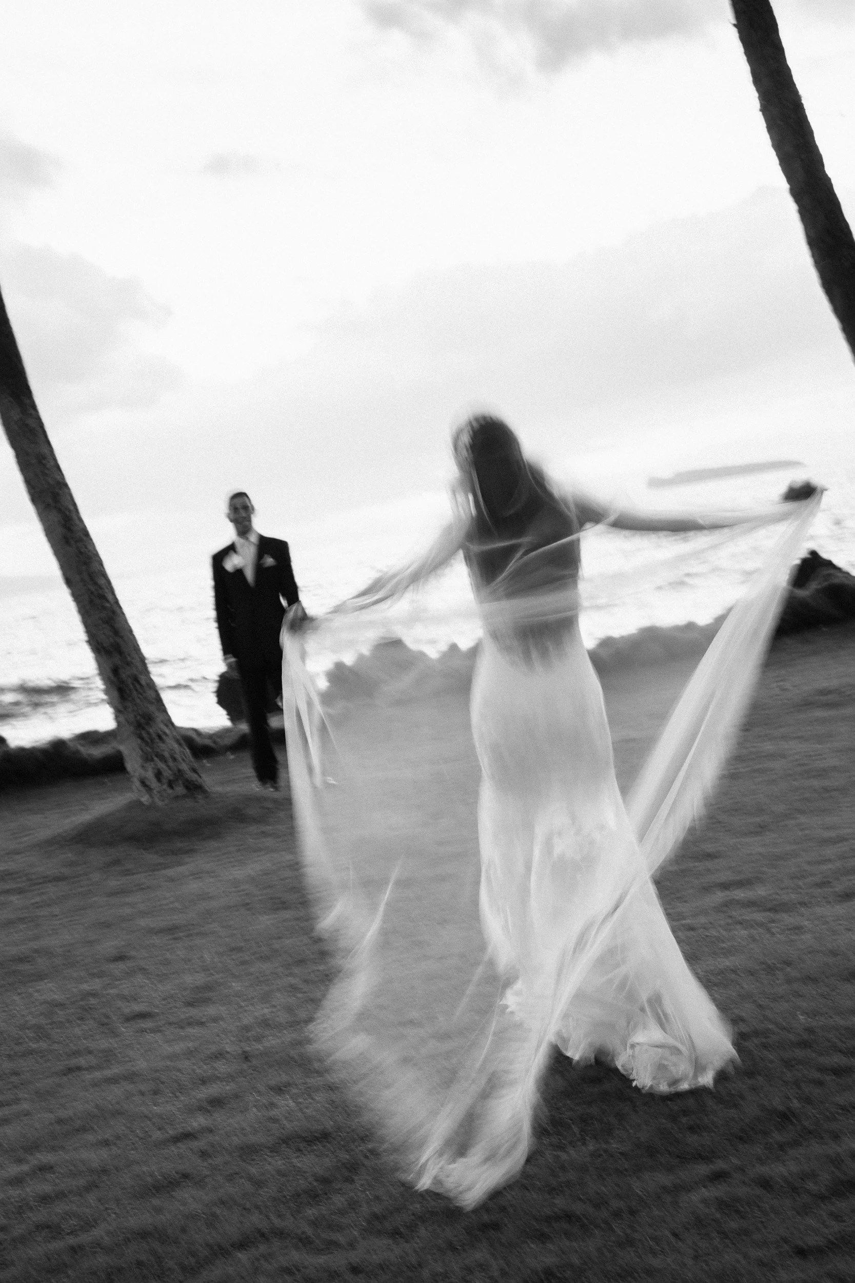 A bride in a wedding dress with a long veil dancing on a beach with an ocean and cloudy sky in the background, a man in a suit watching her.