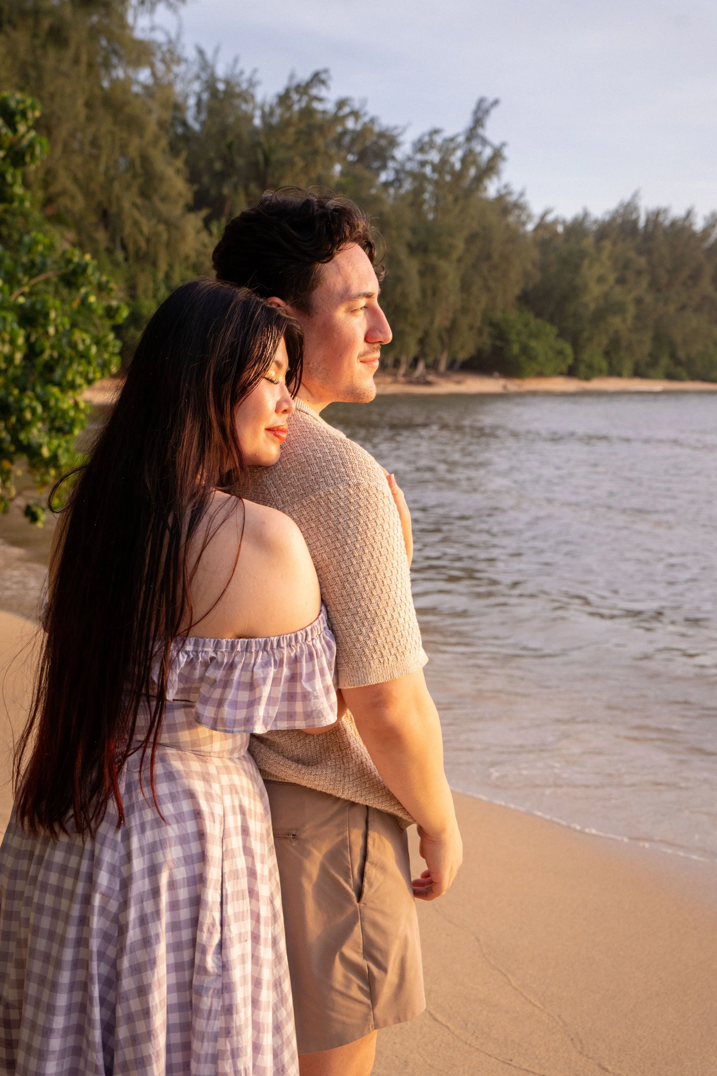 A young woman and young man standing on a sandy beach near the water, with trees in the background, enjoying the sunset with eyes closed.