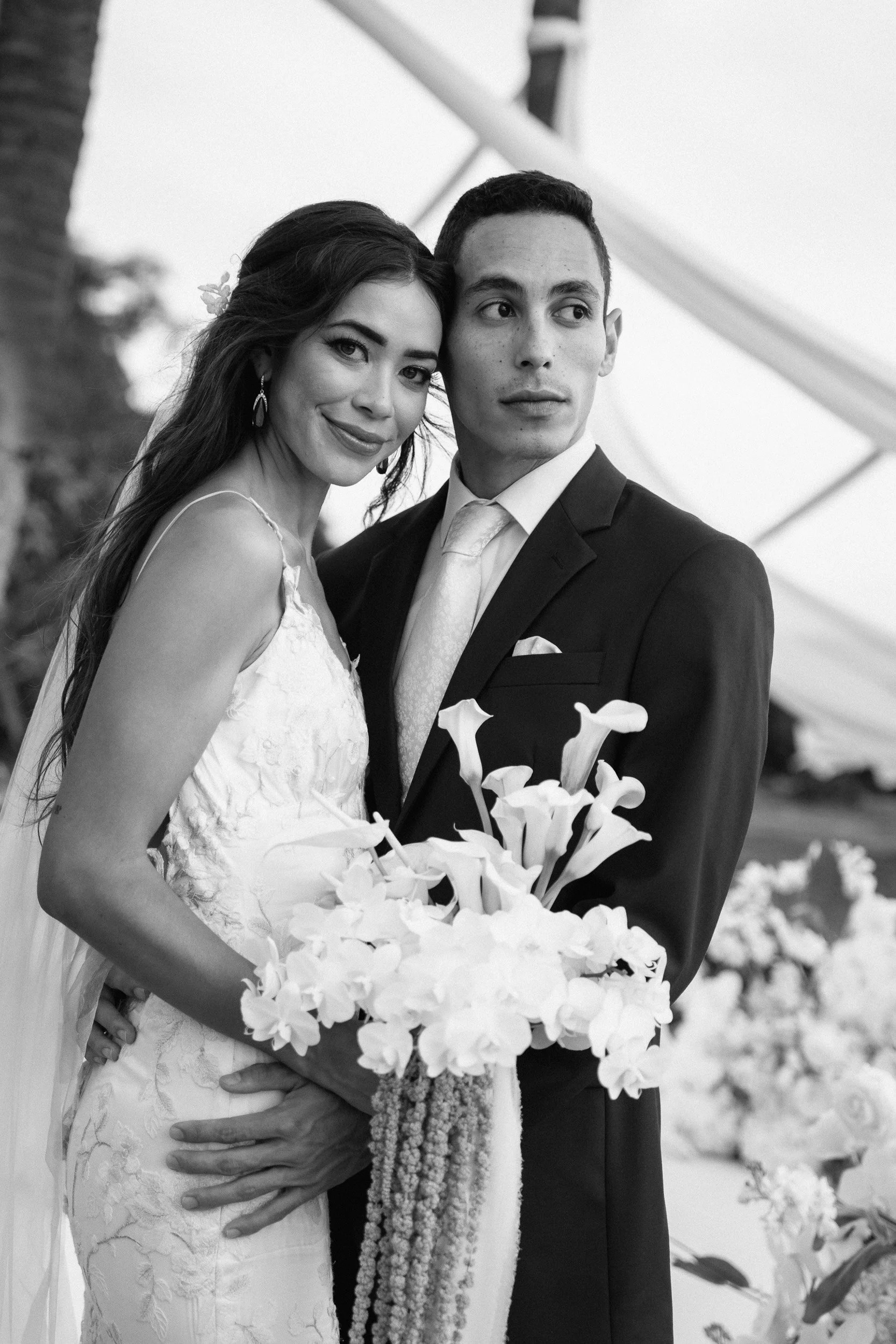 A bride and groom on their wedding day, standing close together and looking to the side. The bride is holding a bouquet of flowers, and they are both dressed in wedding attire.