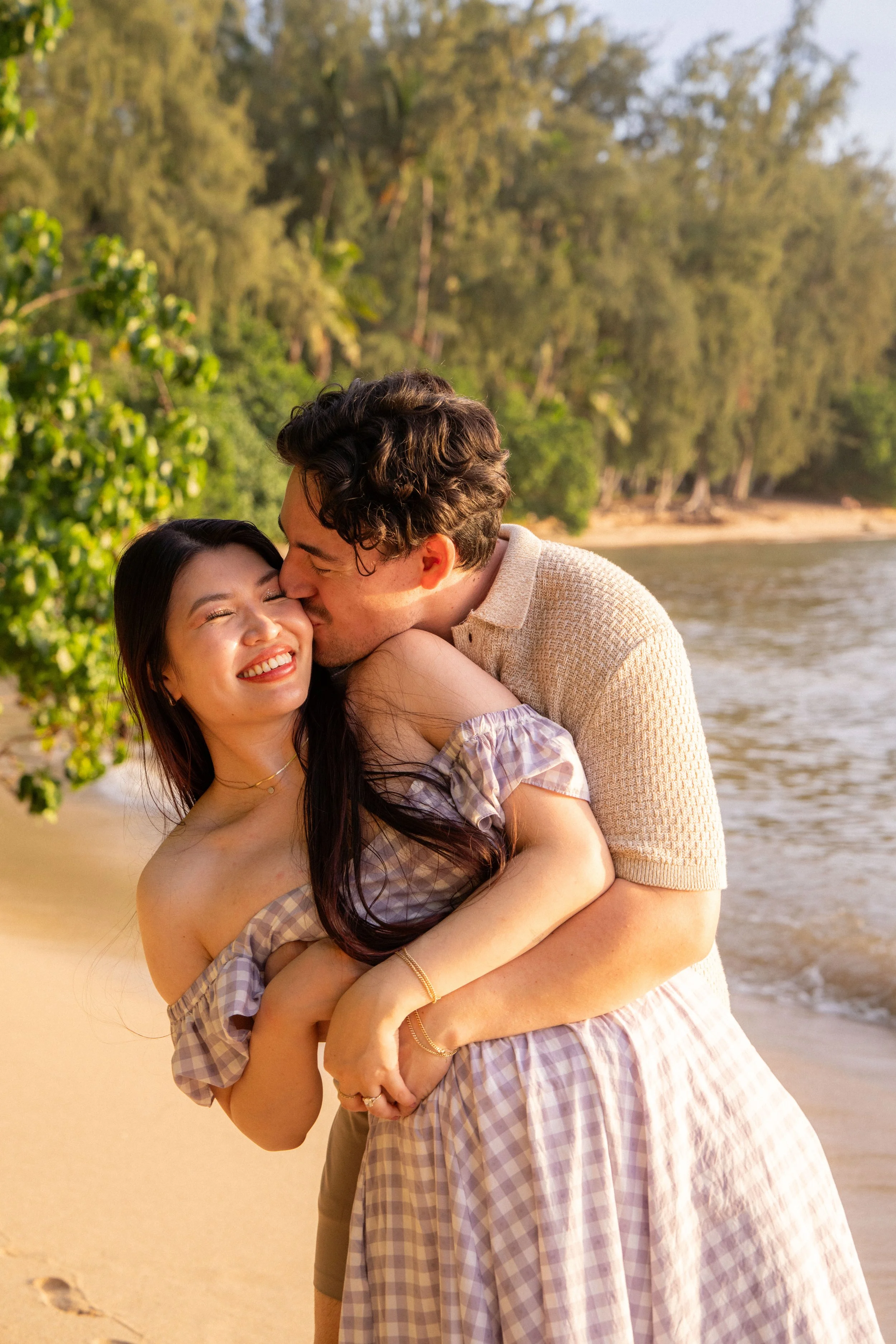 A happy couple hugging by the beach, with trees and water in the background during sunset.