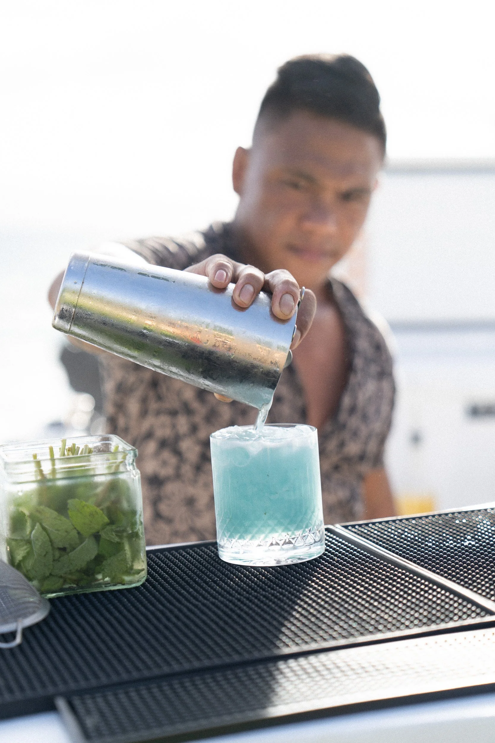Person pouring a blue cocktail into a glass at a bar, with containers of fresh mint and other ingredients on the counter.