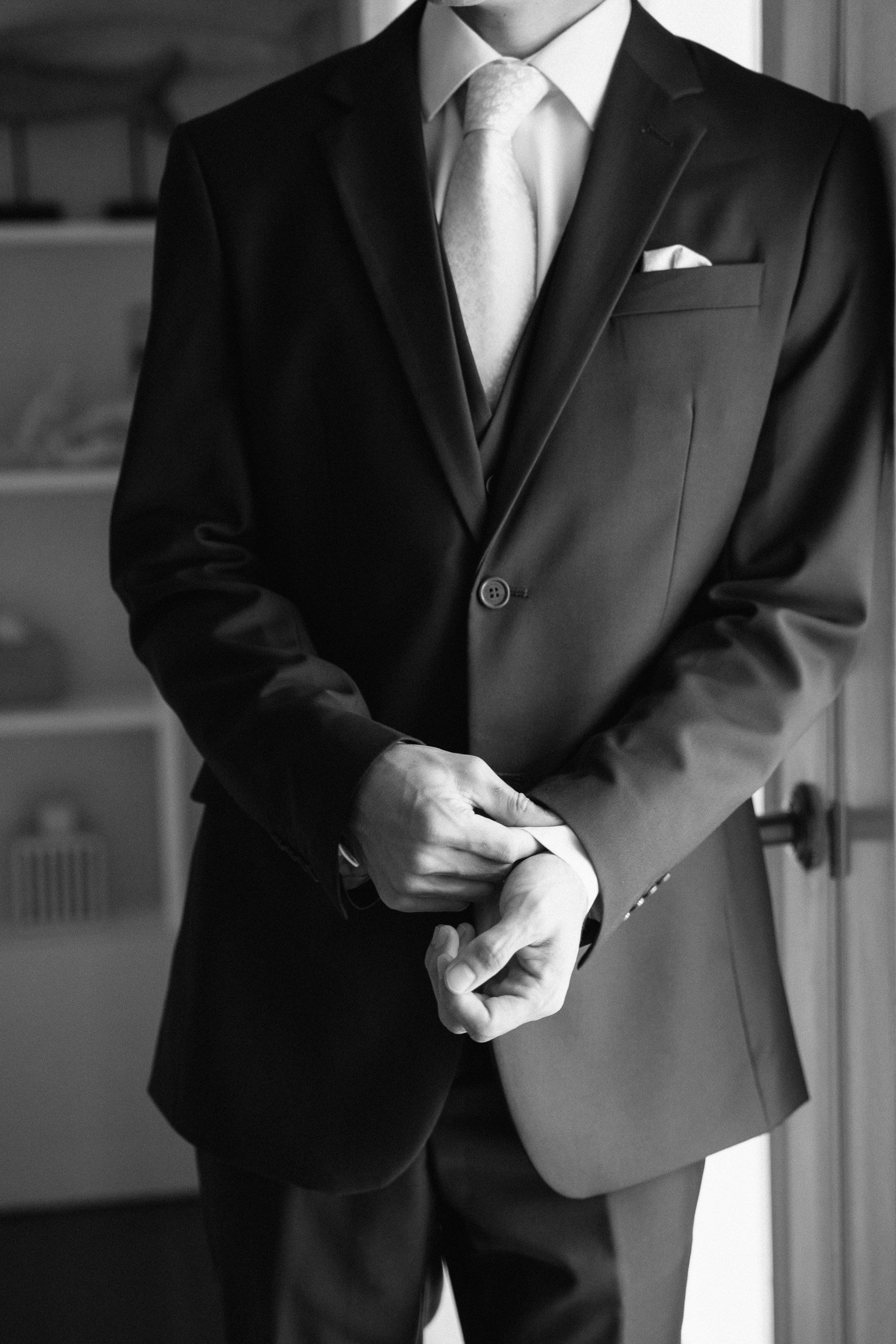 A man in formal attire, wearing a dark suit, white shirt, and tie, adjusting his sleeve in an indoor setting.