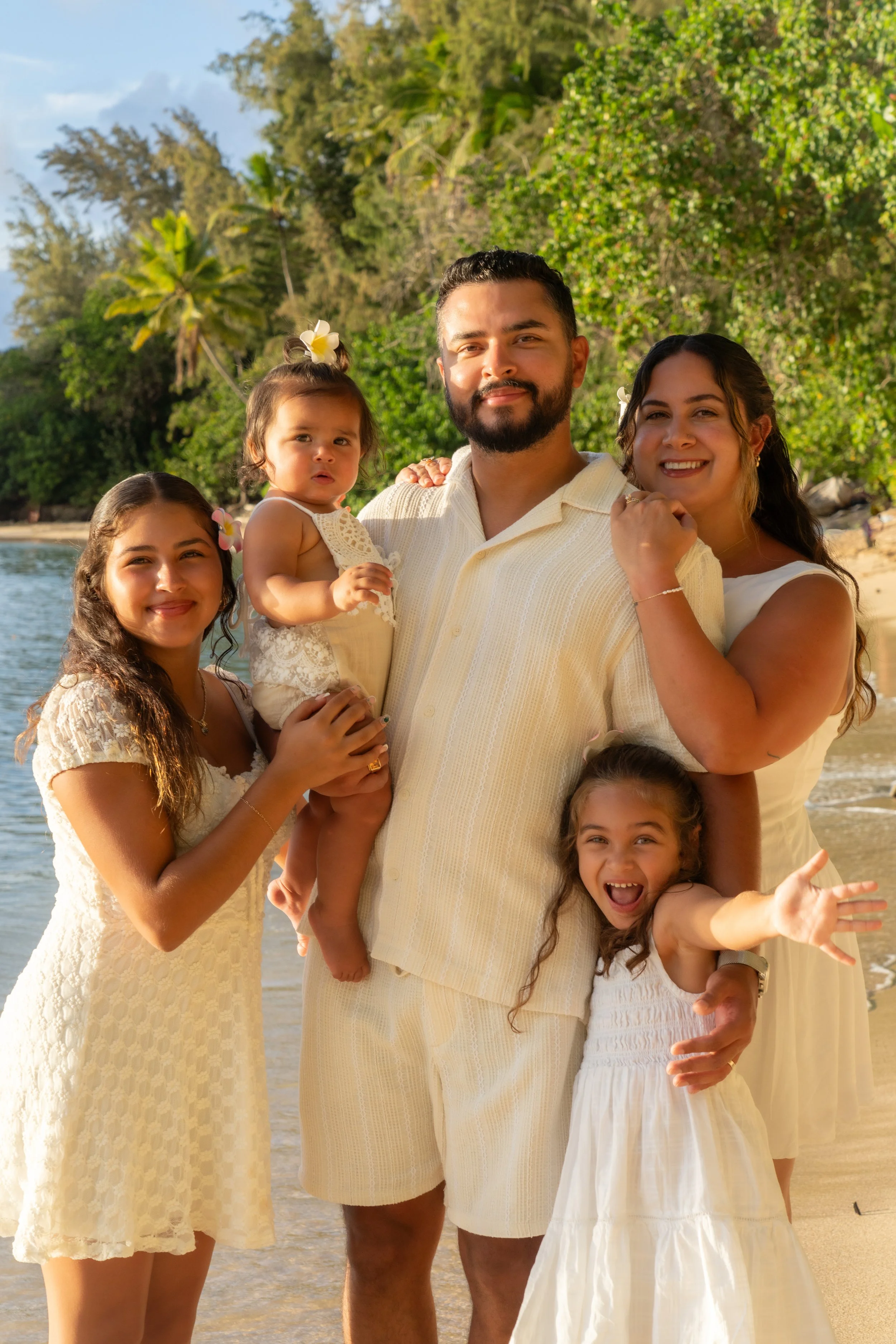 A family of six posing on a beach on the north shore of Oahu, dressed in light-colored summer clothing with greenery and trees in the background.