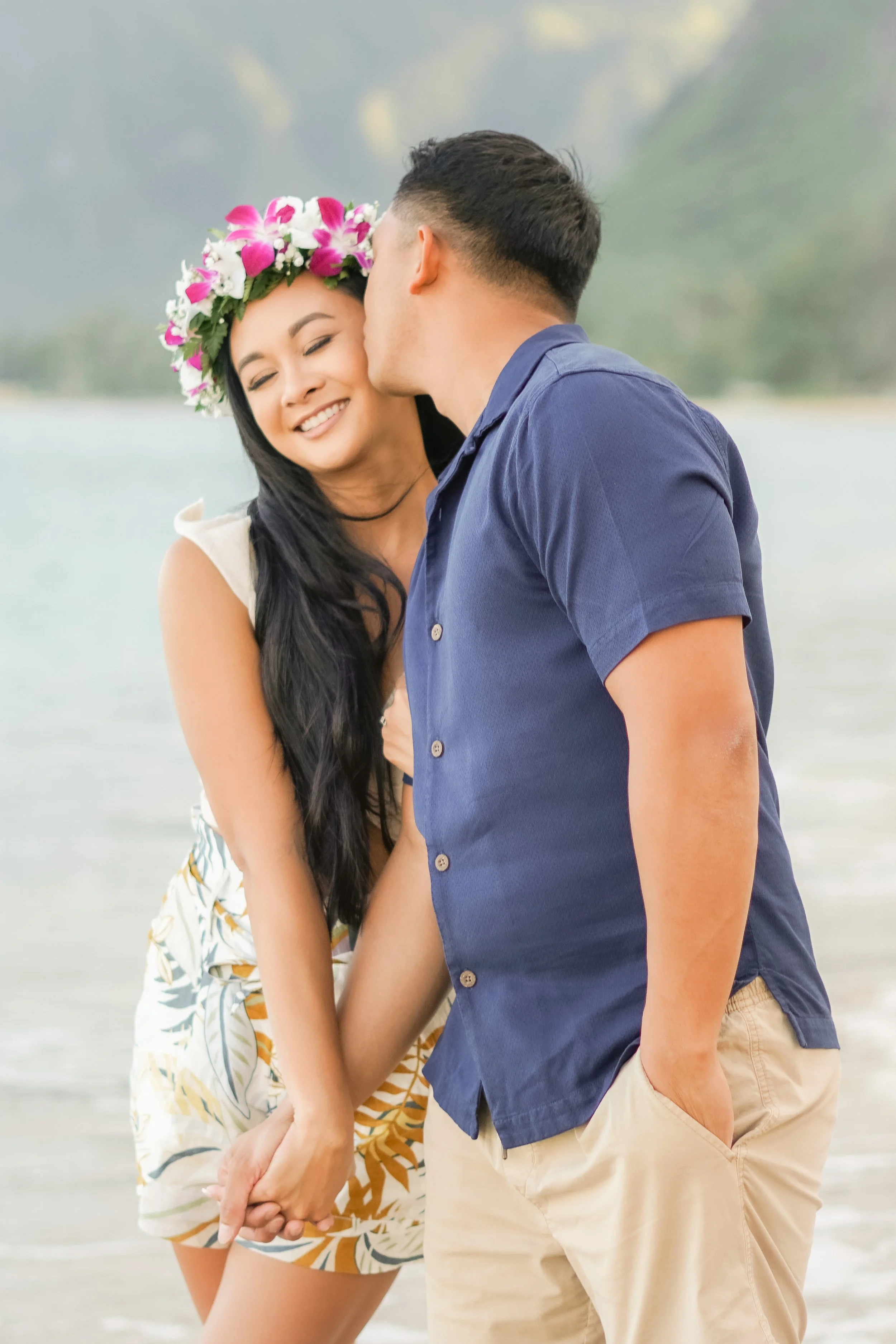 A young couple sharing a kiss while holding hands on the beach, on the east side of oahu, with the woman wearing a floral crown and the man in a blue shirt and khaki shorts.